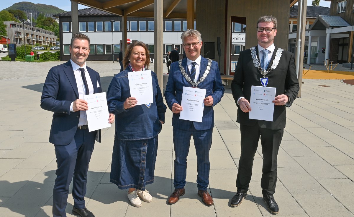 Four individuals standing outdoors, each holding a certificate, with two wearing ceremonial chains, smiling at the camera.