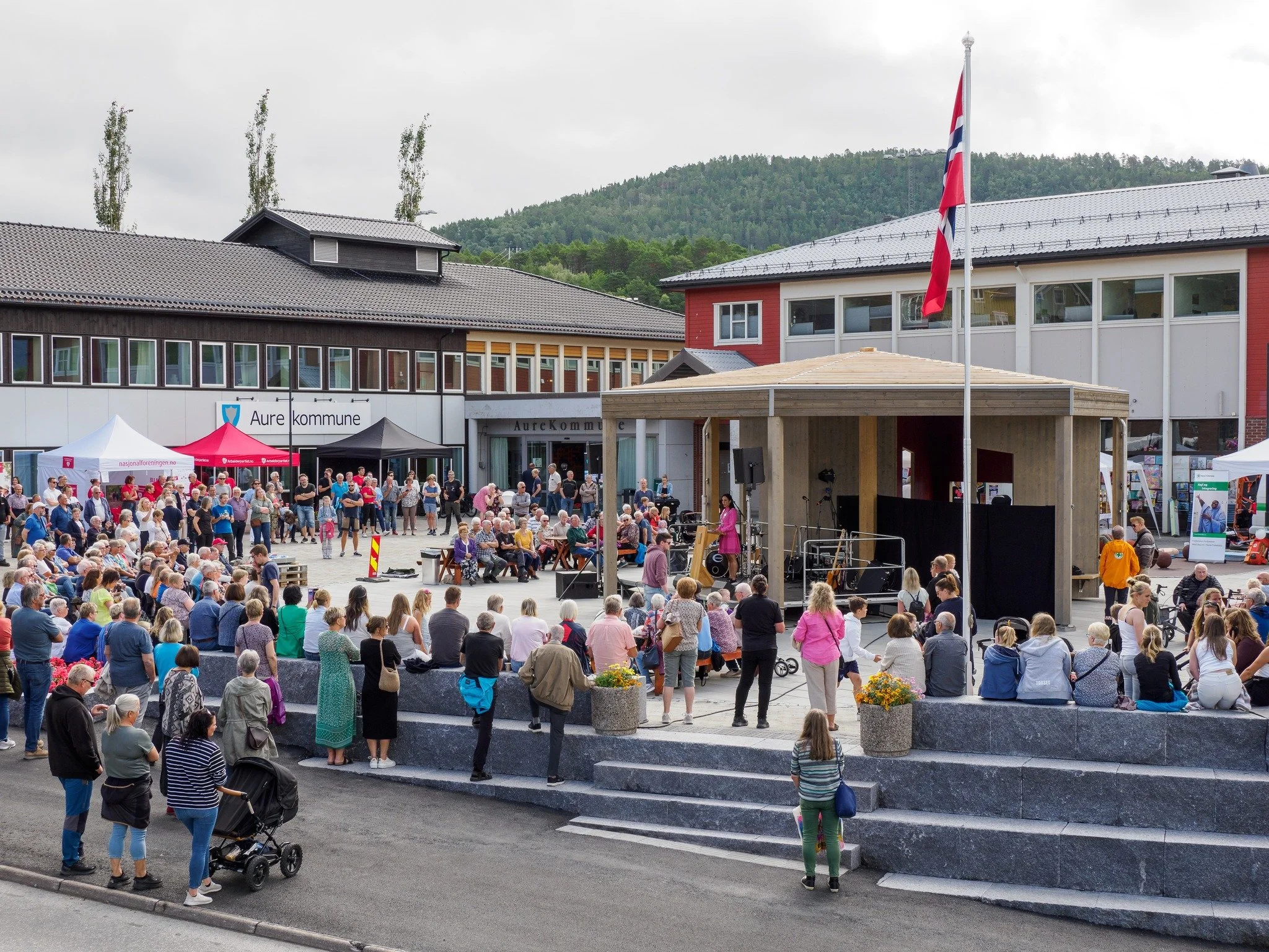 A crowd gathered in front of a public building with the sign 'Aure kommune' during an outdoor event, with tents, a stage, and a Norwegian flag, against a backdrop of mountains and cloudy sky.