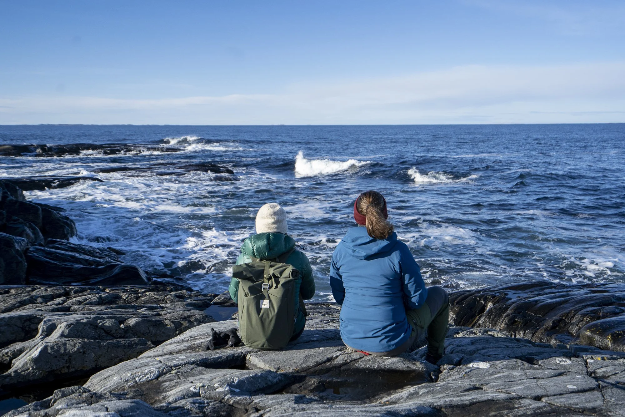 Two people sit on rocky shoreline looking out at the ocean with waves and a blue sky.