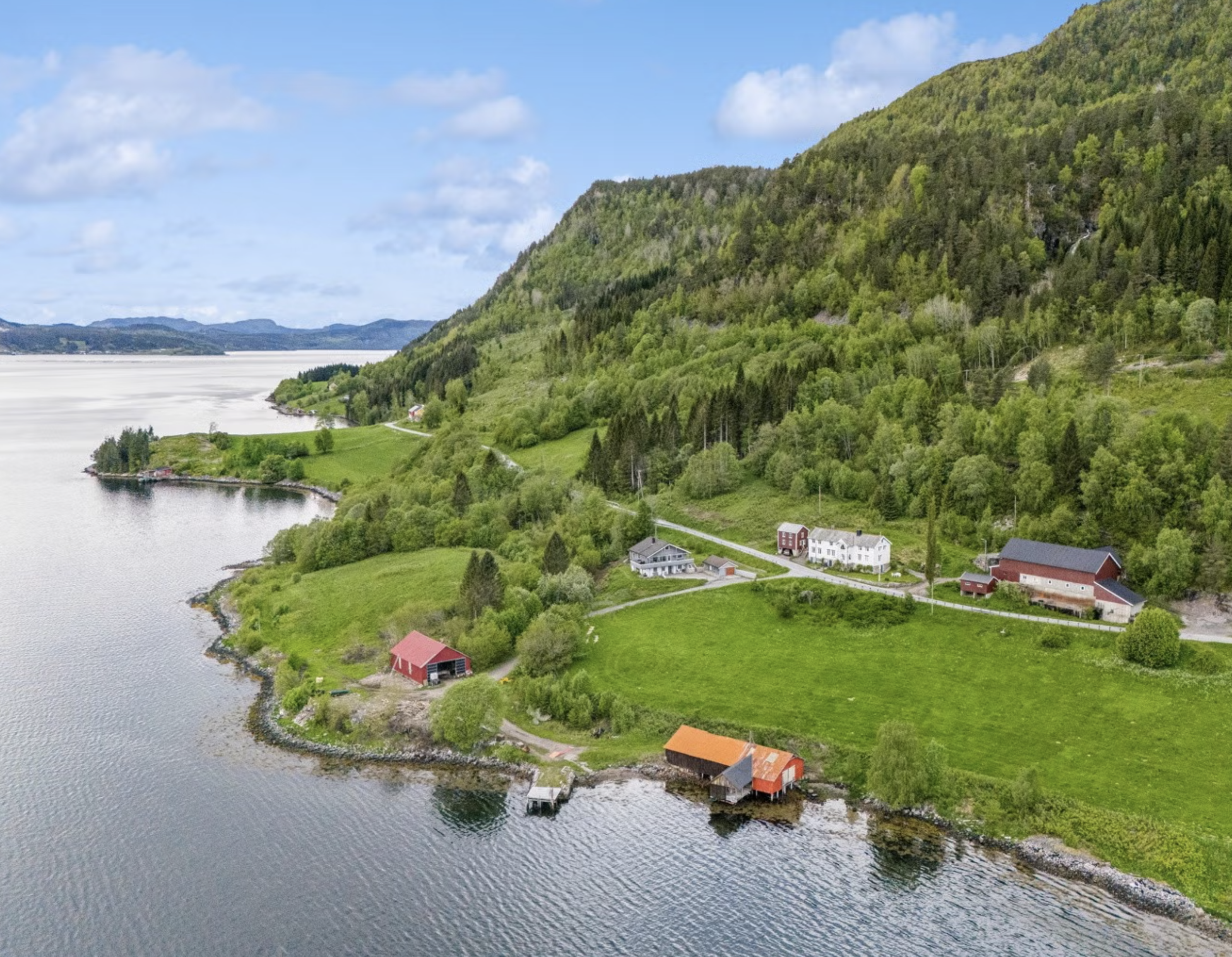 A scenic view of a body of water next to lush green hills and trees, with several houses and barns along the shoreline under a blue sky with scattered clouds.