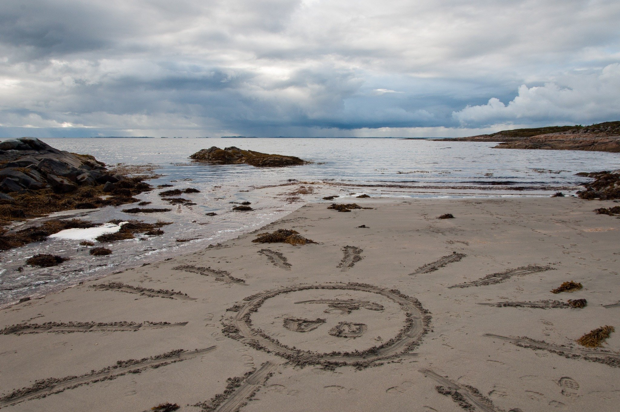 A sandy beach with tracks and seaweed, rocks on the shore, calm water, and a cloudy sky.