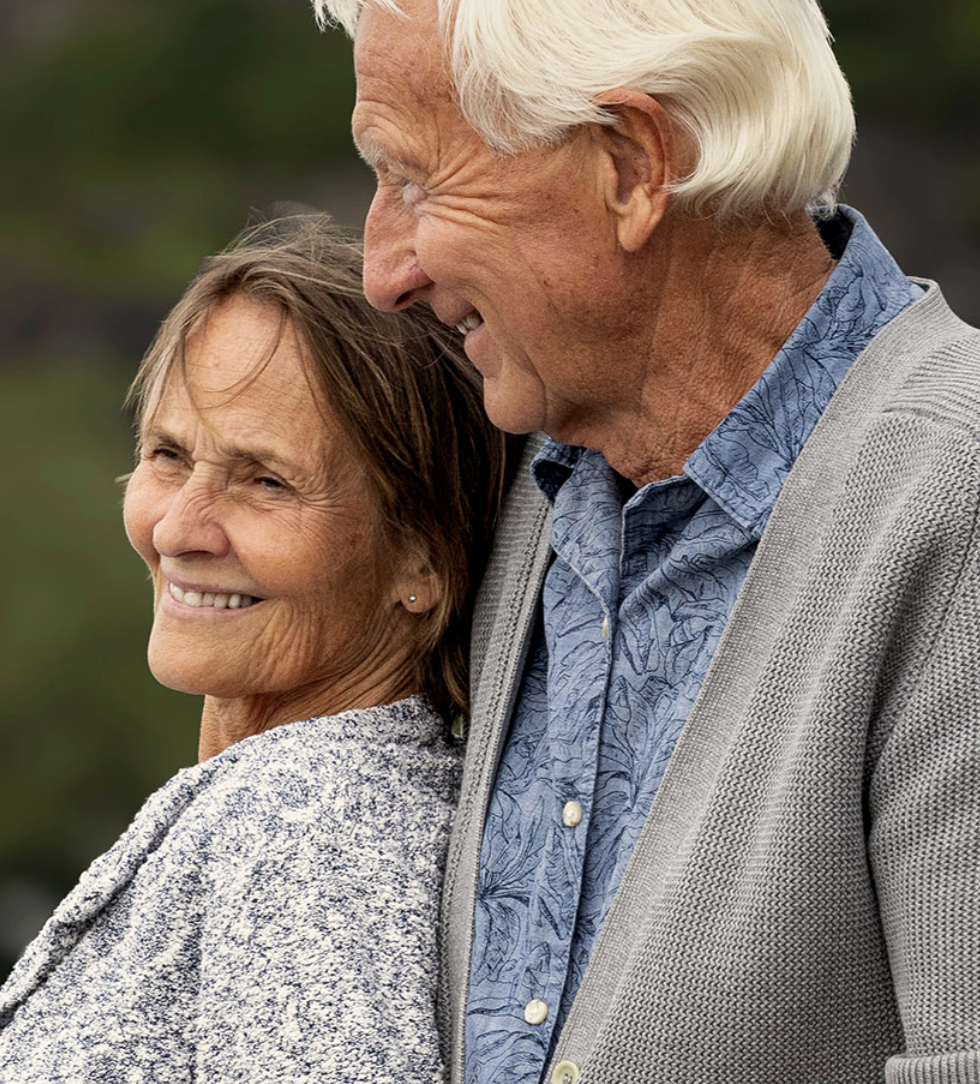 An elderly man and woman smiling and looking at each other outdoors, with the man wearing a blue patterned shirt and gray cardigan, and the woman wearing a textured gray sweater.