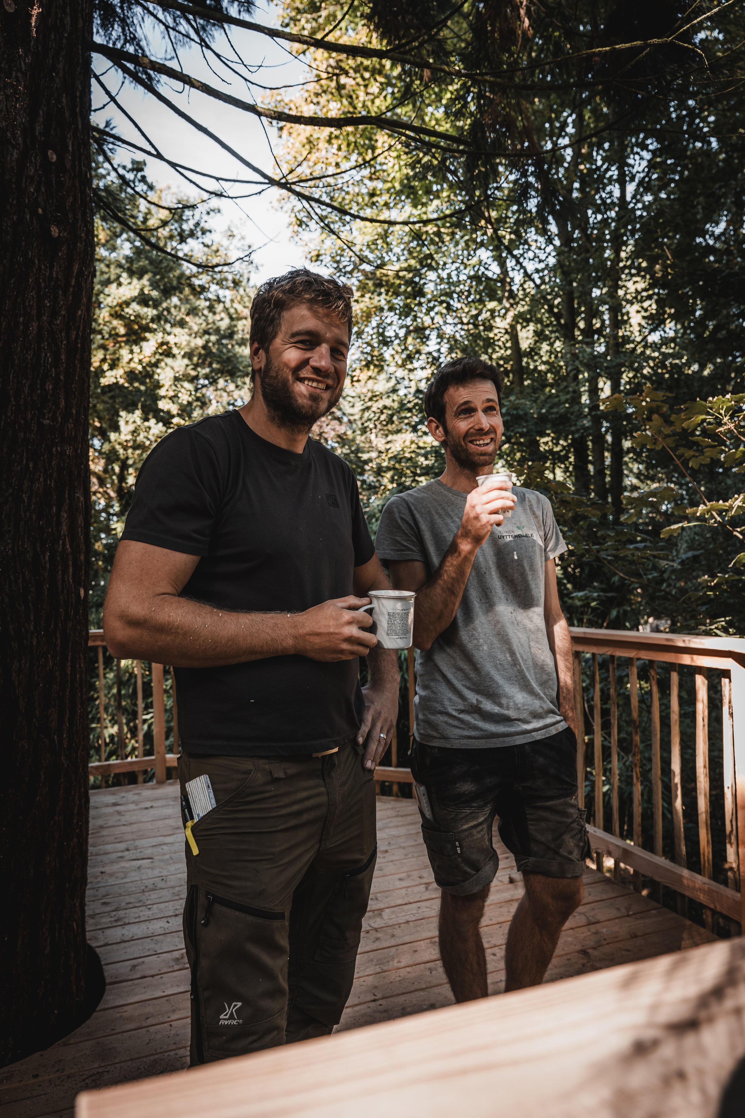 Twee mannen staan ​​op een houten vlonder in een weelderig bos, met kopjes in hun handen en een glimlach op hun gezicht.
