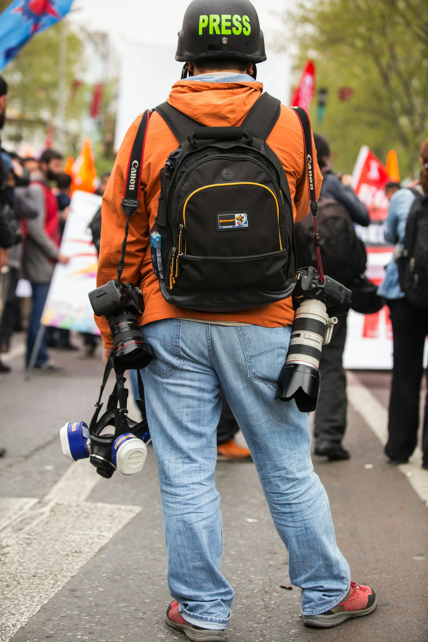 Un homme portant un casque avec la inscription 'PRESS', un sac à dos, une caméra avec long objectif, une caméra avec masque respiratoire, un autre appareil, en arrière-plan une foule manifestante avec des pancartes et drapeaux.