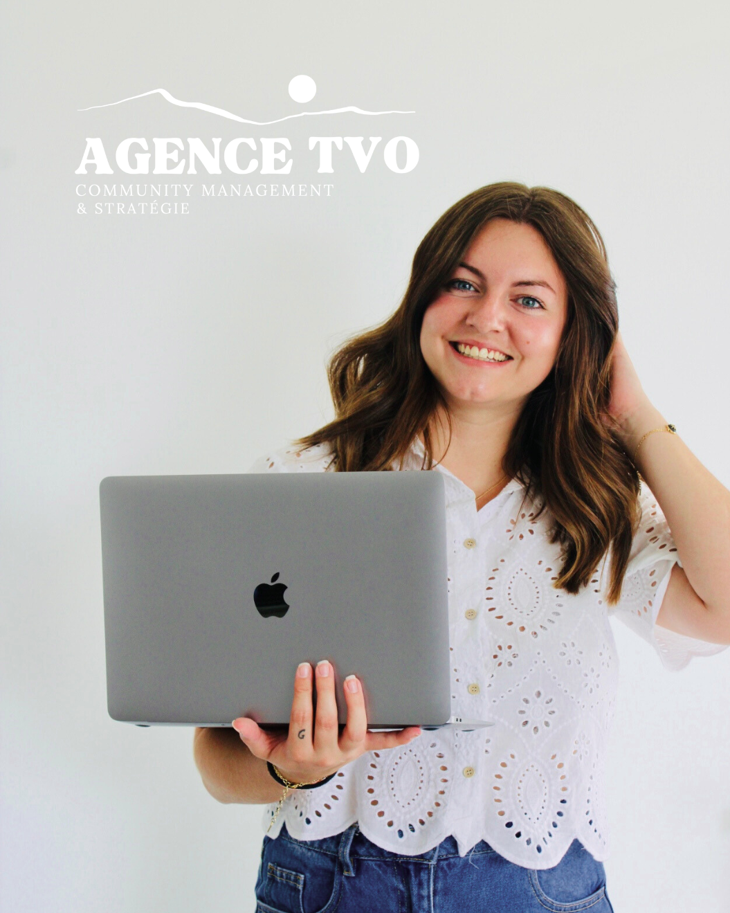 Une femme souriante tenant un ordinateur portable devant un mur blanc avec le logo d'une agence de communication.