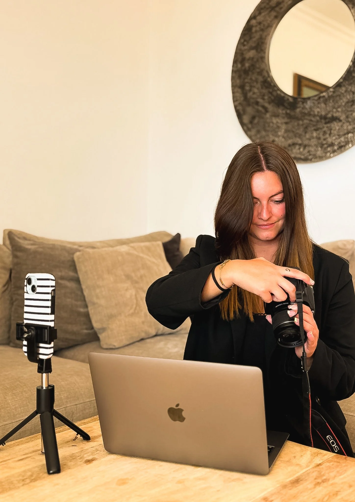 Une femme prépare une vidéo ou une séance photo avec un téléphone fixe sur une petite tripie, un ordinateur portable, et une caméra, dans un salon avec un grand miroir rond et un canapé avec des coussins.