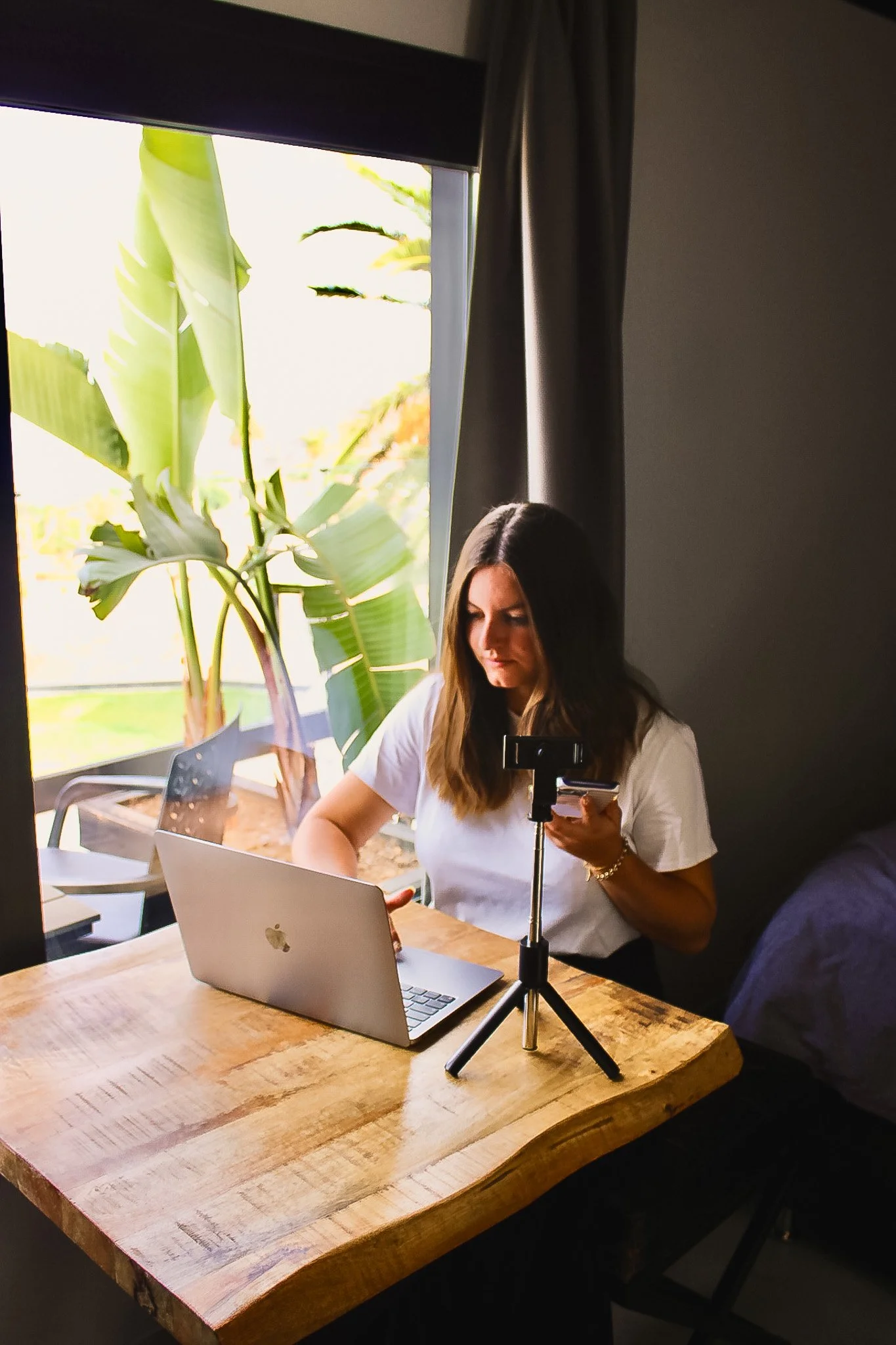 Une femme assise à une table en bois, utilisant un ordinateur portable Apple, avec un smartphone et un trépied posés devant elle, près d'une fenêtre avec une grande plante verte à l'extérieur.