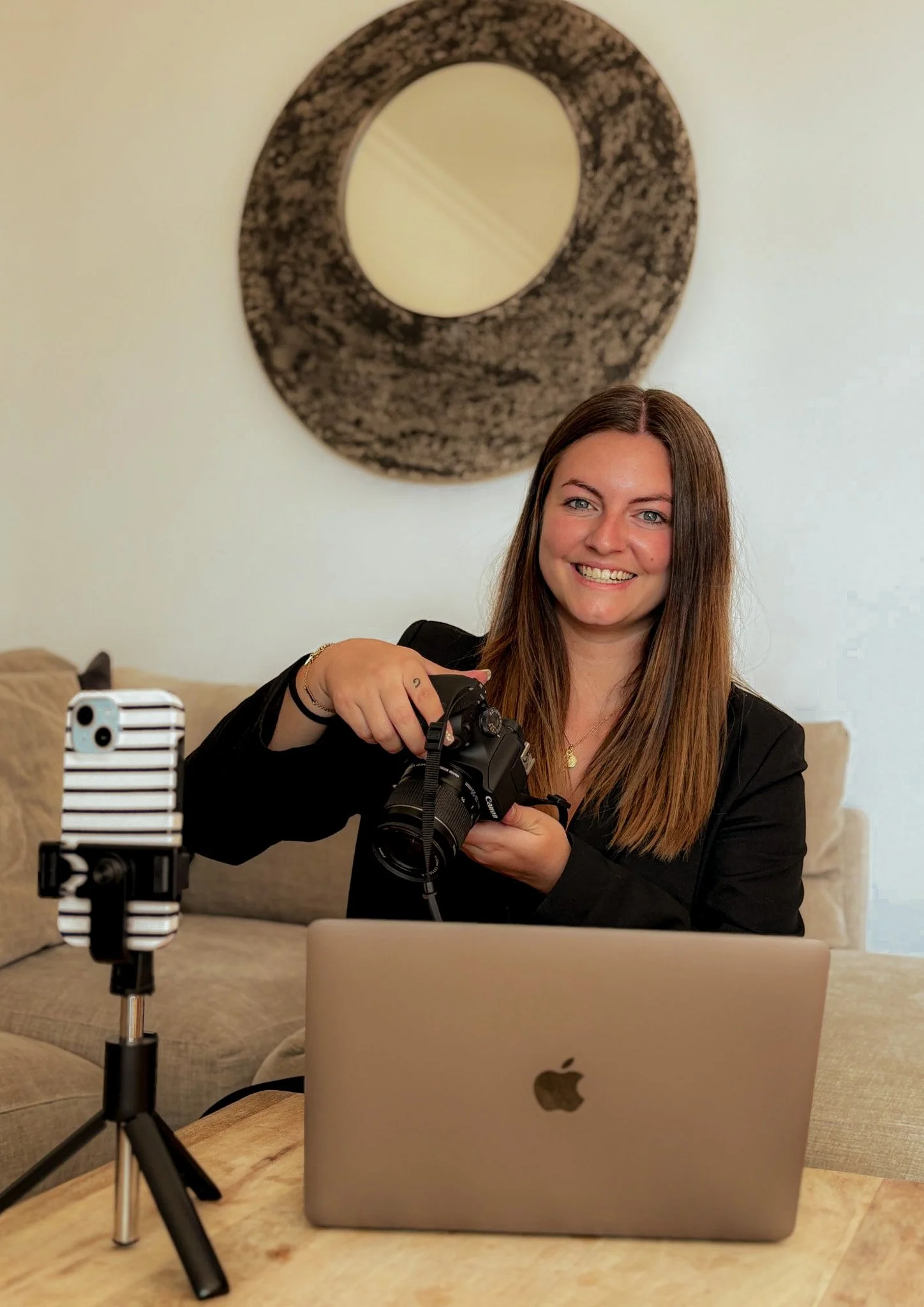 Une femme souriante tient un appareil photo en main, assise à une table avec un ordinateur portable Apple. Un téléphone est monté sur un trépied à gauche, dans un intérieur avec un canapé et un tableau abstrait noir et blanc au mur.