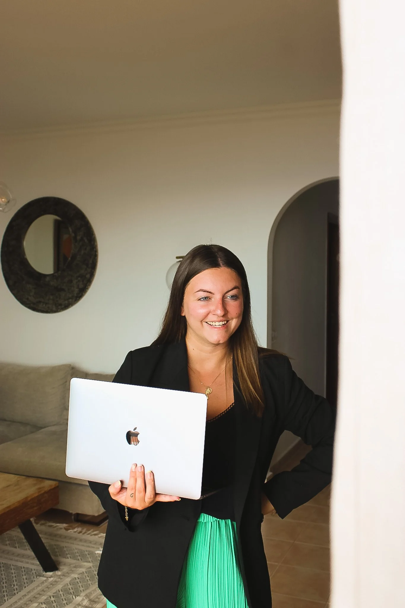Une femme souriante regarde un ordinateur portable blanc dans un intérieur domestique moderne.