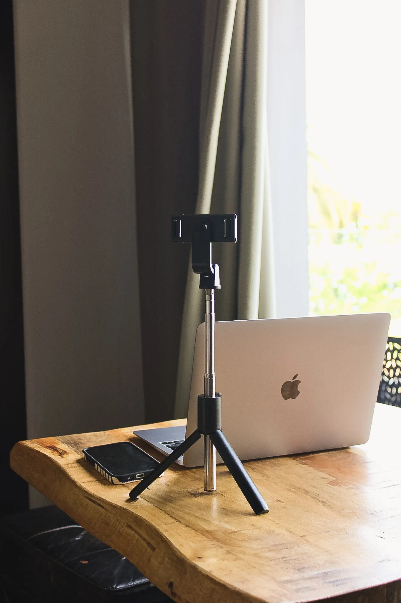 Table en bois avec un MacBook, un téléphone portable et un support pour smartphone ou tablette, près d'une fenêtre avec rideaux