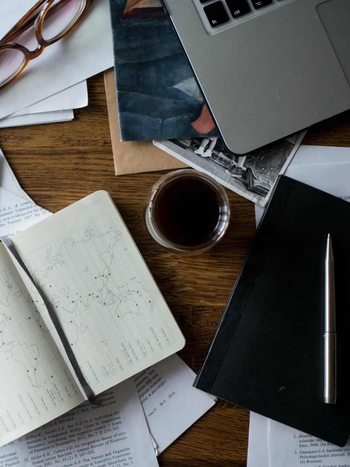 Ein Arbeitsraum mit mehreren Notizbüchern, einer Tasse Kaffee, einem Laptop, einem schwarzen Buch mit Kugelschreiber, Zeitungspapieren und eine Brille auf einem Holztisch.