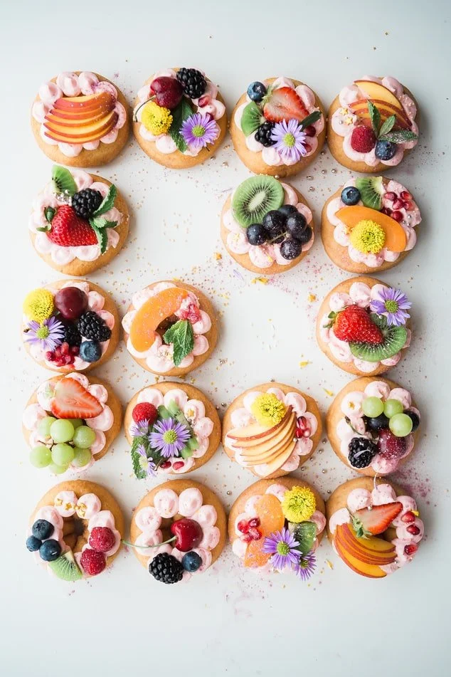 Viele kleine Törtchen mit buntem Obst, Blumen und Creme dekoriert, auf weißem Hintergrund