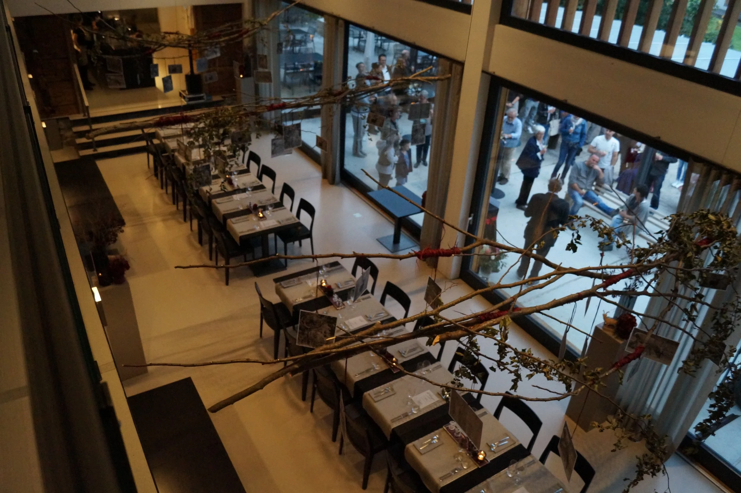 Leerer Speisesaal mit gedeckten Tischen, große Fenster mit Blick nach draußen, draußen versammelte Menschen, eine dekorative Astgarnitur in der Mitte des Raumes.