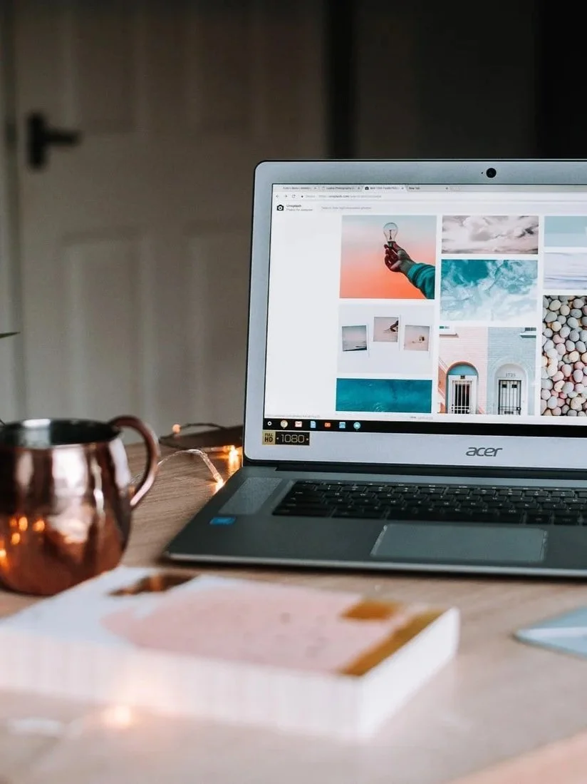 Ein Laptop auf einem Holzschreibtisch, daneben eine kupferfarbene Tasse und eine Schachtel mit Keksen. Der Bildschirm zeigt eine Webseite mit Fotos.