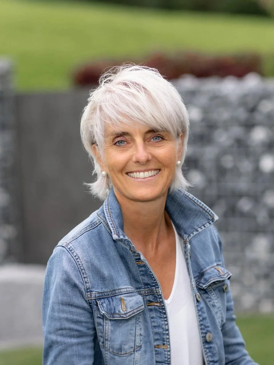 Eine lachende ältere Frau mit kurzen, weißen Haaren, trägt eine blaue Jeansjacke und weiße Perlenohrringe steht im Freien vor einem grauen Steinmauer und grünem Hintergrund.