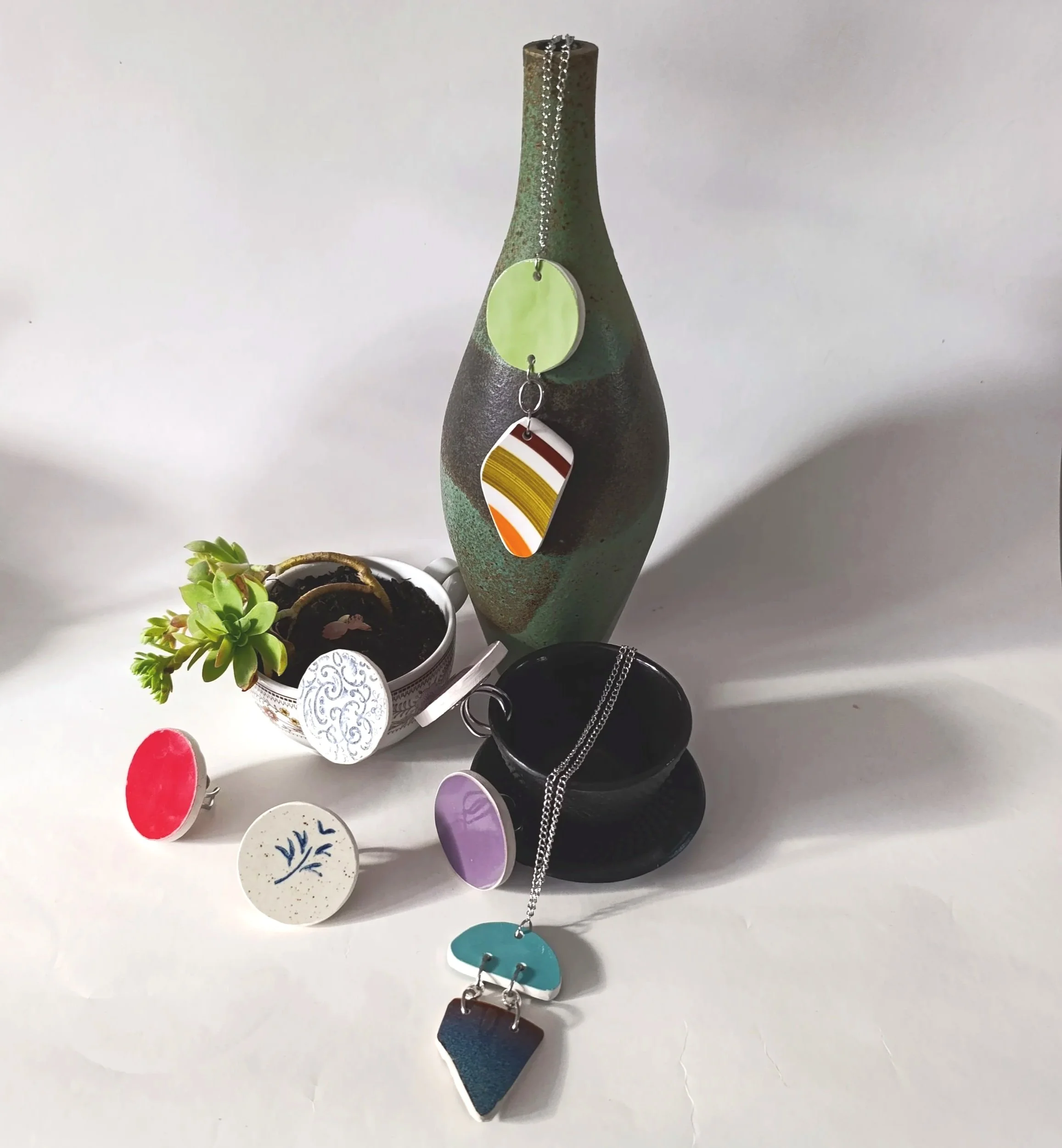 Vase en céramique ou en terre cuite contenant une petite succulente, plusieurs bijoux en céramique upcyclés, dont colliers, bagues avec des designs colorés, et un bracelet, disposés sur une surface blanche.