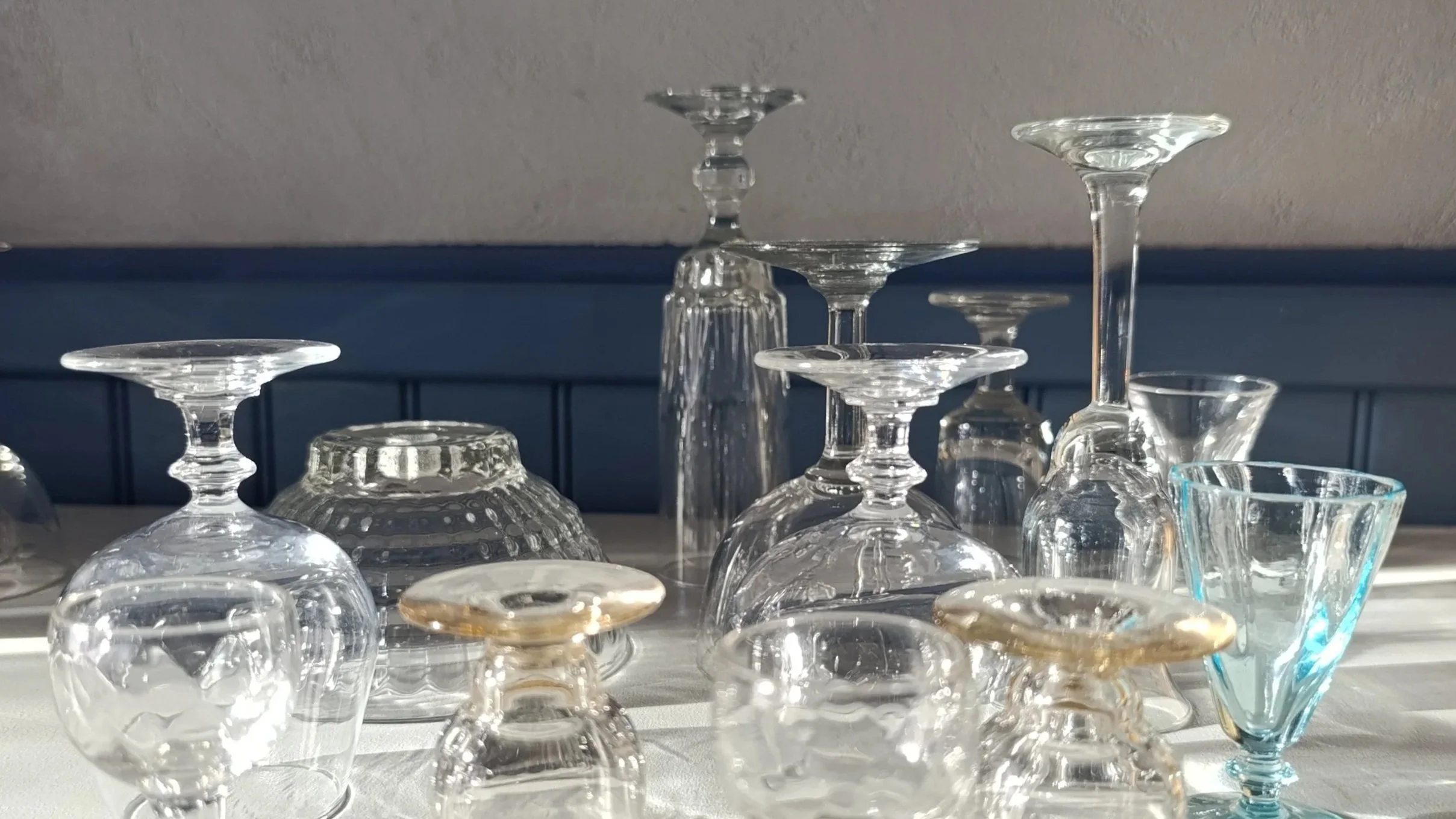 Verres en verre sur une surface ensoleillée, certains sont à l'envers, d'autres droits, avec un fond de mur gris et une plinthe bleue.