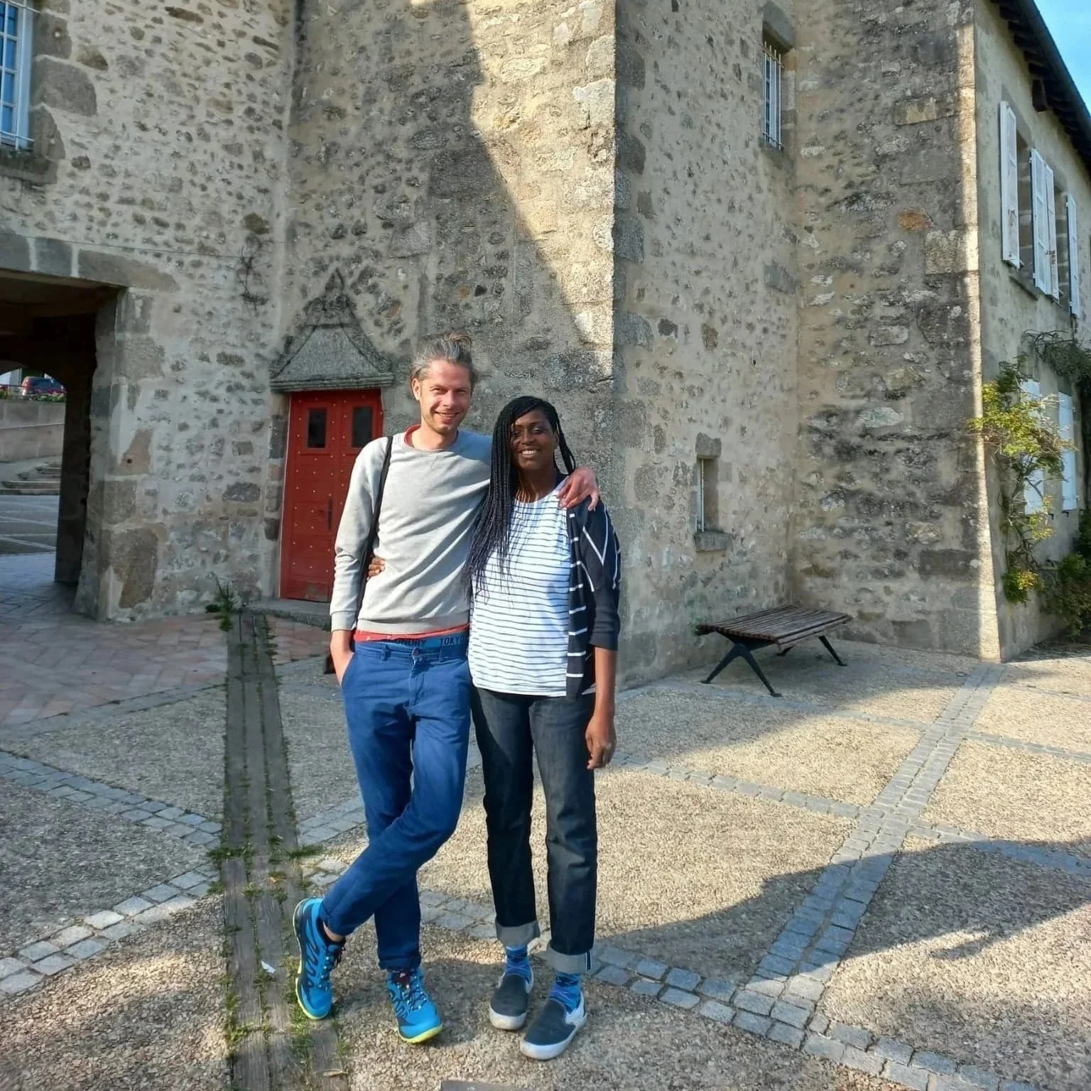 Deux personnes souriantes, un homme et une femme, posent ensemble devant une vieille bâtisse en pierre, à l'extérieur.