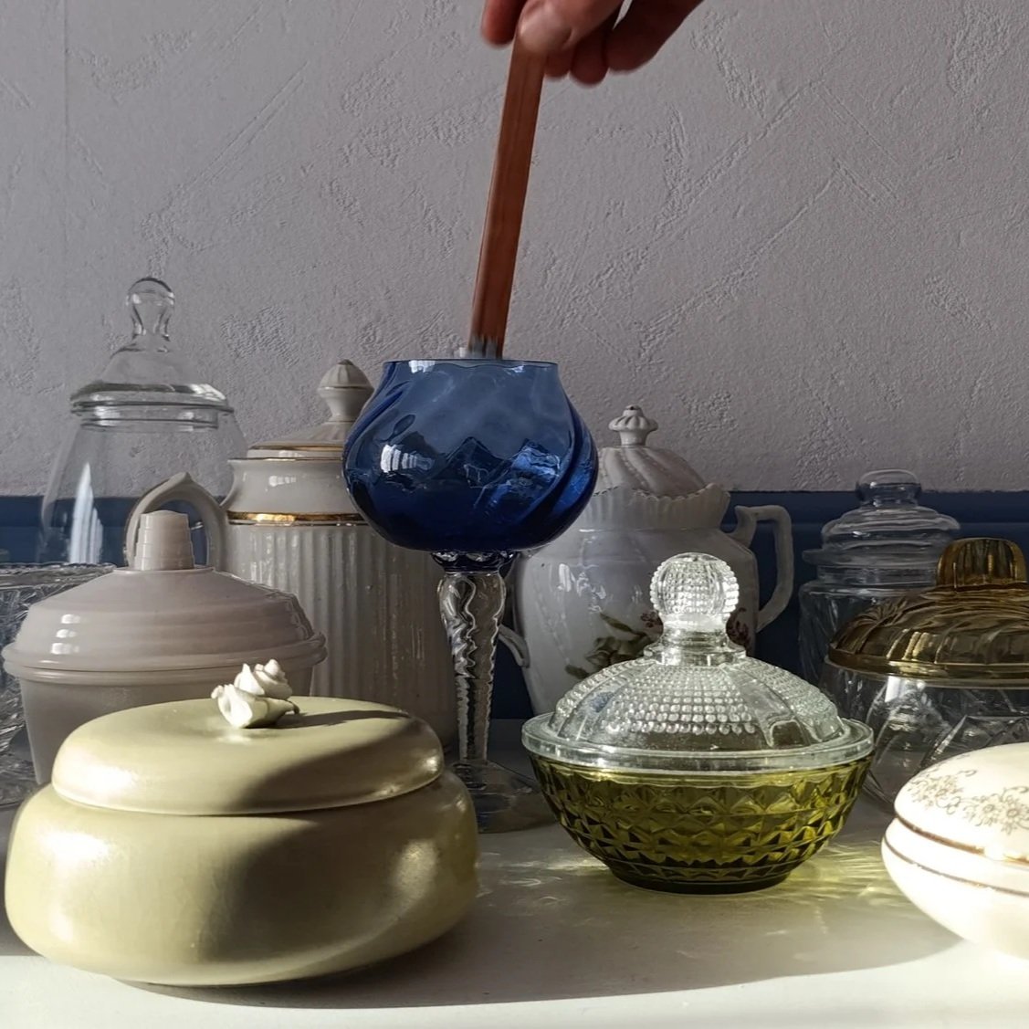 Vue de plusieurs bocaux et pots en verre sur une surface, certains avec couvercles, certains sans, avec un fond blanc et un mur gris en arrière-plan. Un main tient une baguette en bois au-dessus d'un vase en verre bleu.