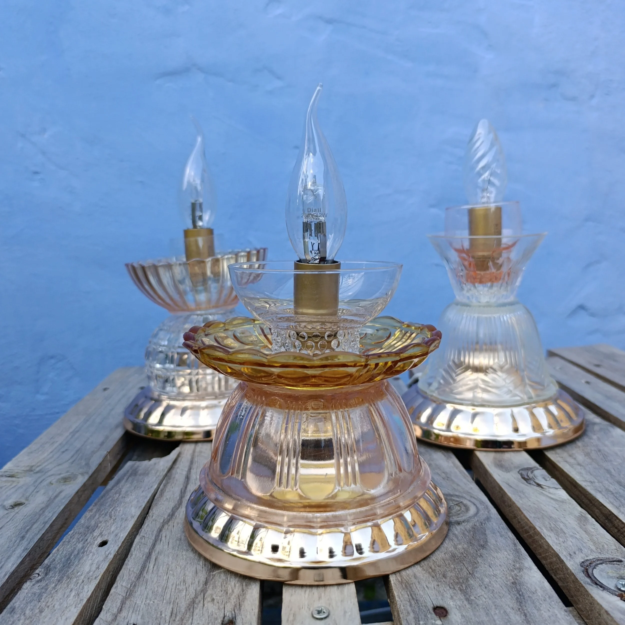 Trois lampes à huile en verre coloré avec formes ondulées, posées sur une surface en bois, contre un mur bleu.