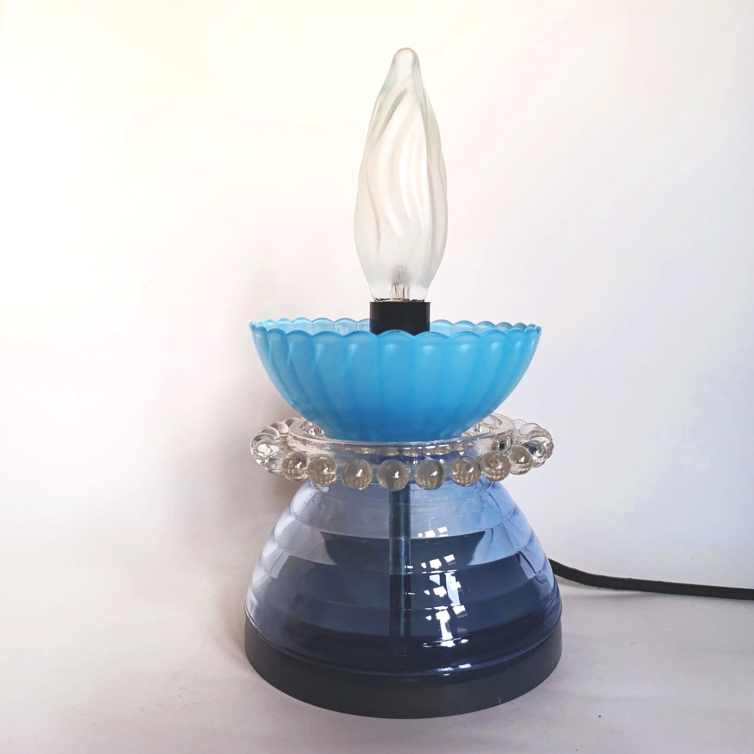Lampe de chevet avec base en verre, coupelle bleue, décoration en perles en verre, et ampoule en forme de flamme.