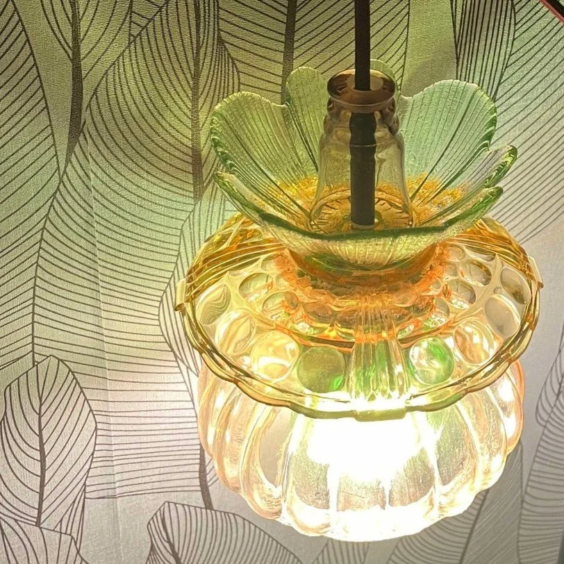 Lampe de plafond en verre teinté avec motif floral, éclairée, sur un fond de papier peint à motifs de feuilles.