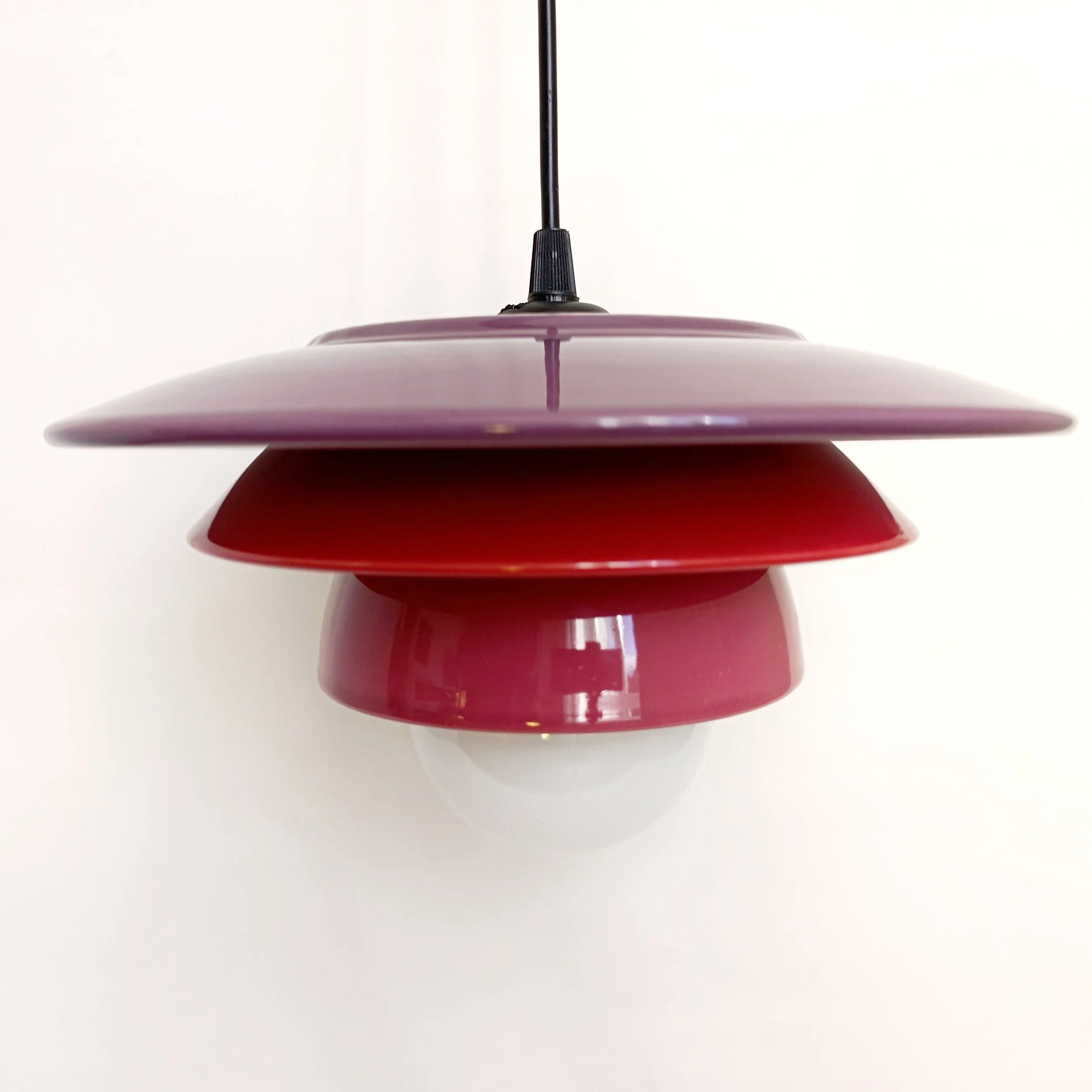Lampe suspendue avec plusieurs disques de couleurs variées, allant du violet, rouge, et rose clair, suspendue par un câble noir, contre un fond blanc