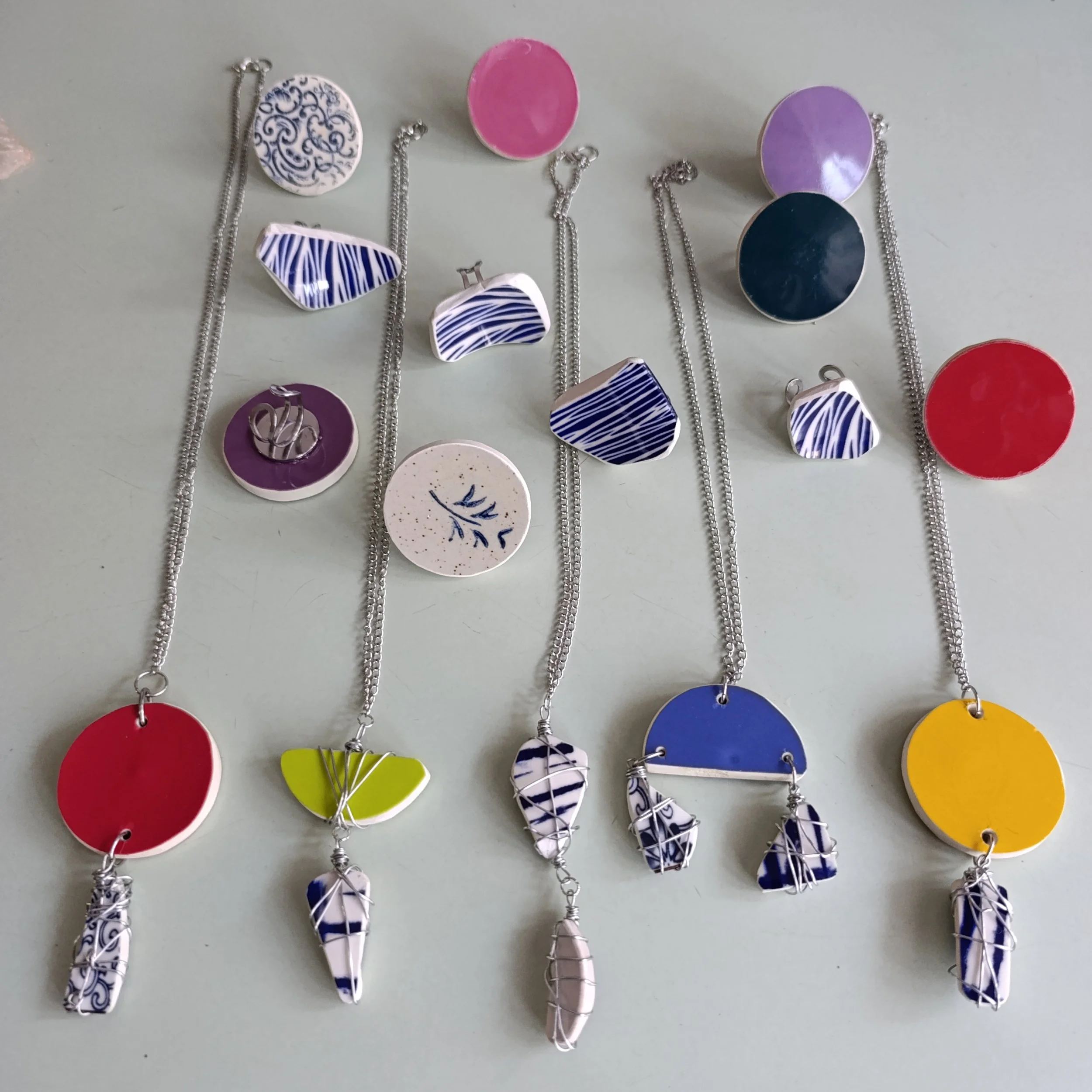 Colliers artisanaux en céramique colorée avec pendentifs en forme de cercles, demi-cercles et formes géométriques, certains ornés de motifs bleus, rouges, jaunes, violets, et blancs, avec des chaînettes en métal argenté.