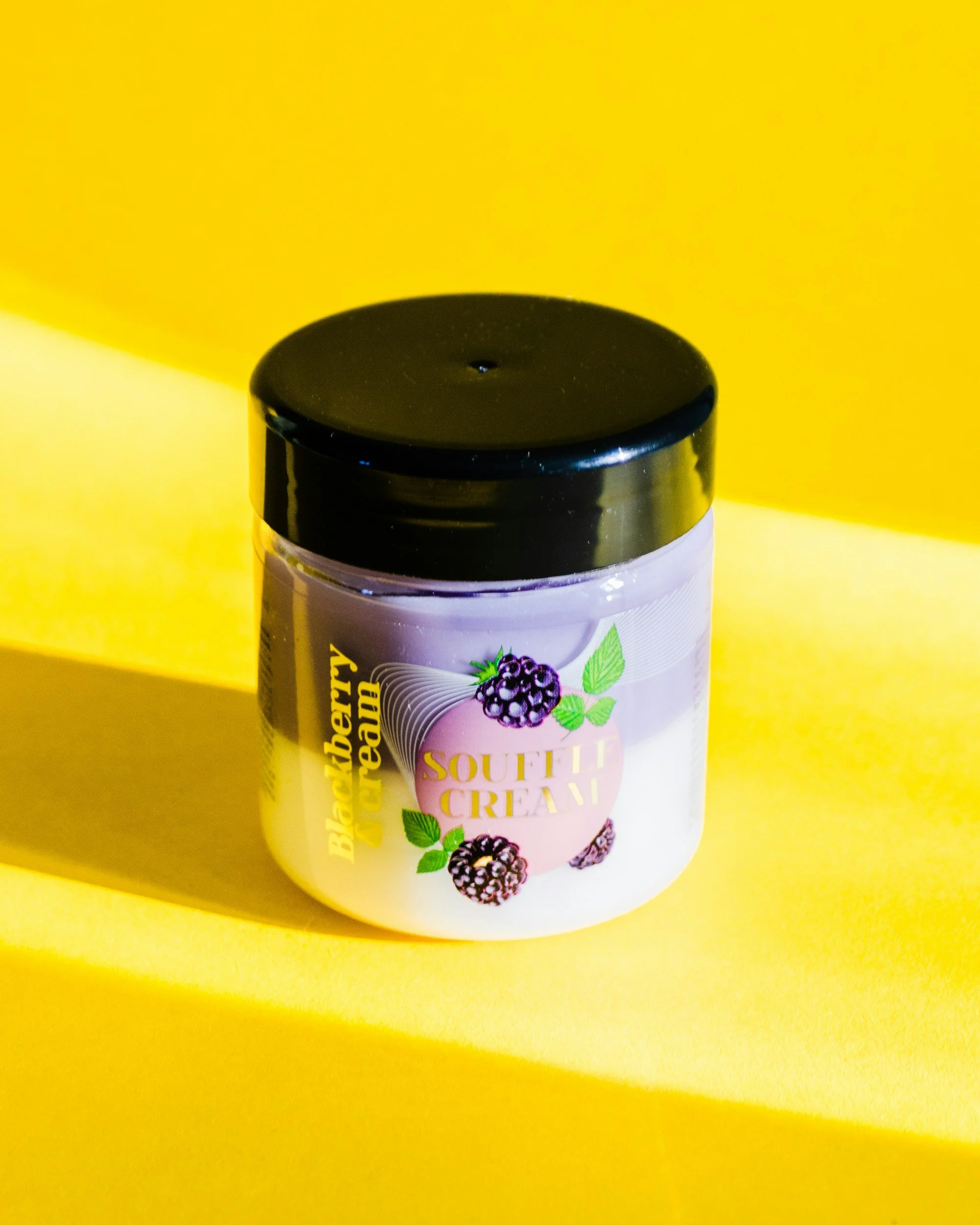 A jar of blackberry and pear flavored souflee cream with a black lid, placed on a yellow background.