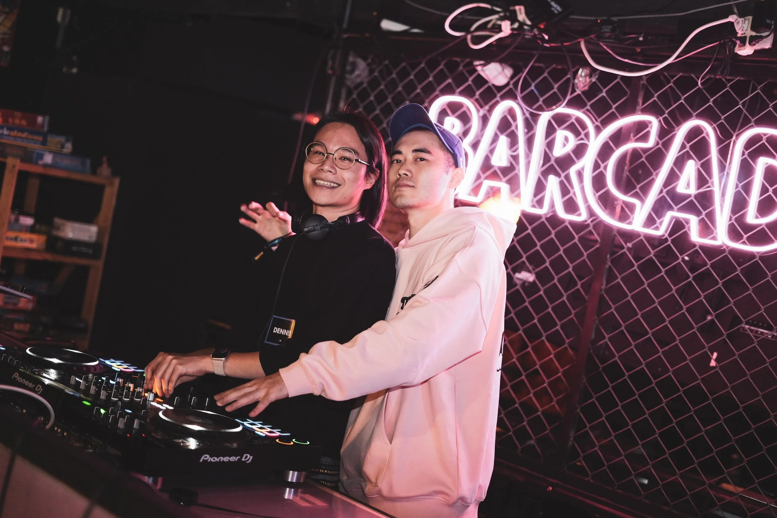 Two young men are DJing in a nightclub with neon pink signs behind them, one smiling and the other looking serious, surrounded by DJ equipment.