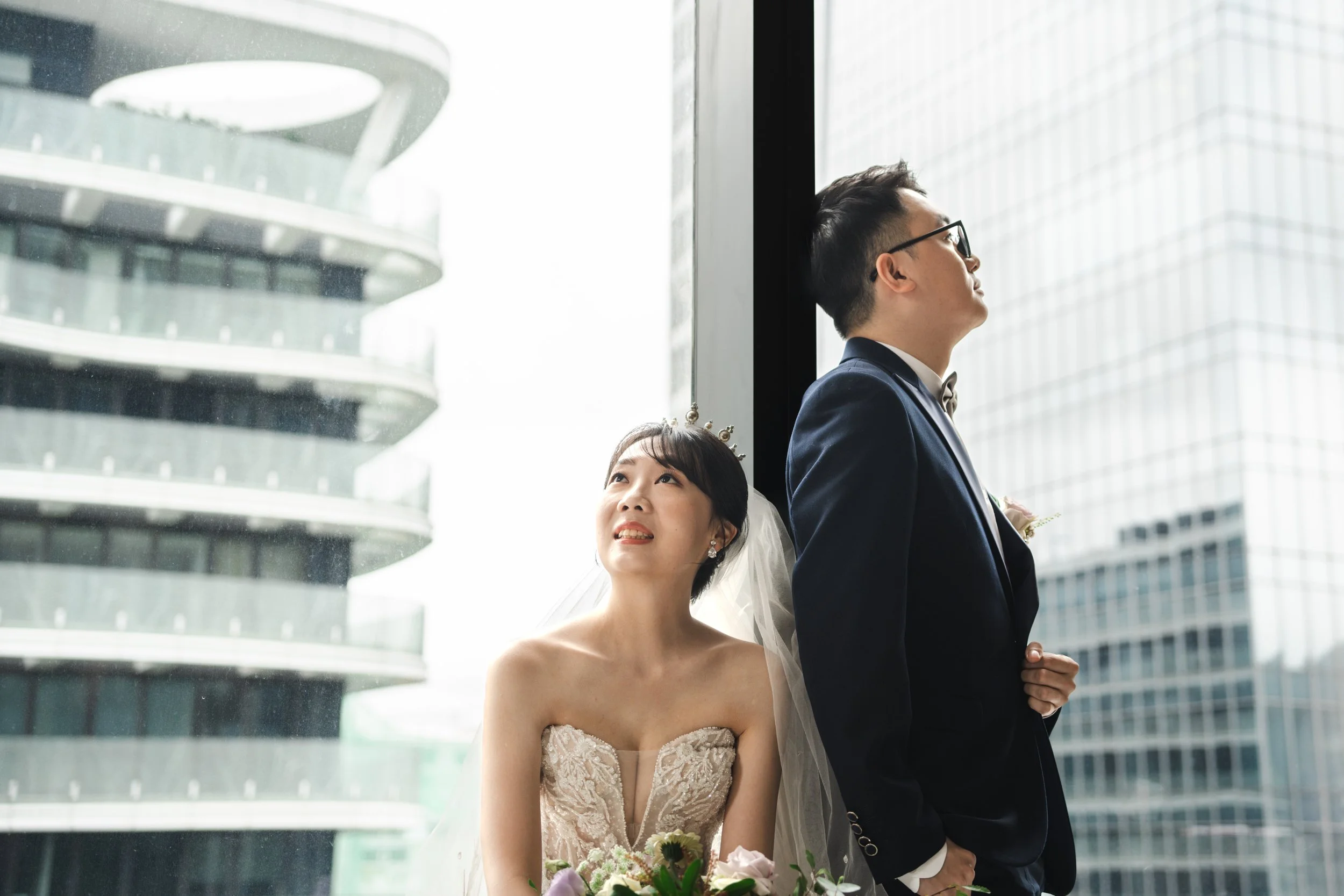 A bride and groom standing indoors near a large window in a modern cityscape, with the bride holding a bouquet and looking up at the groom, who stands with his back to her.