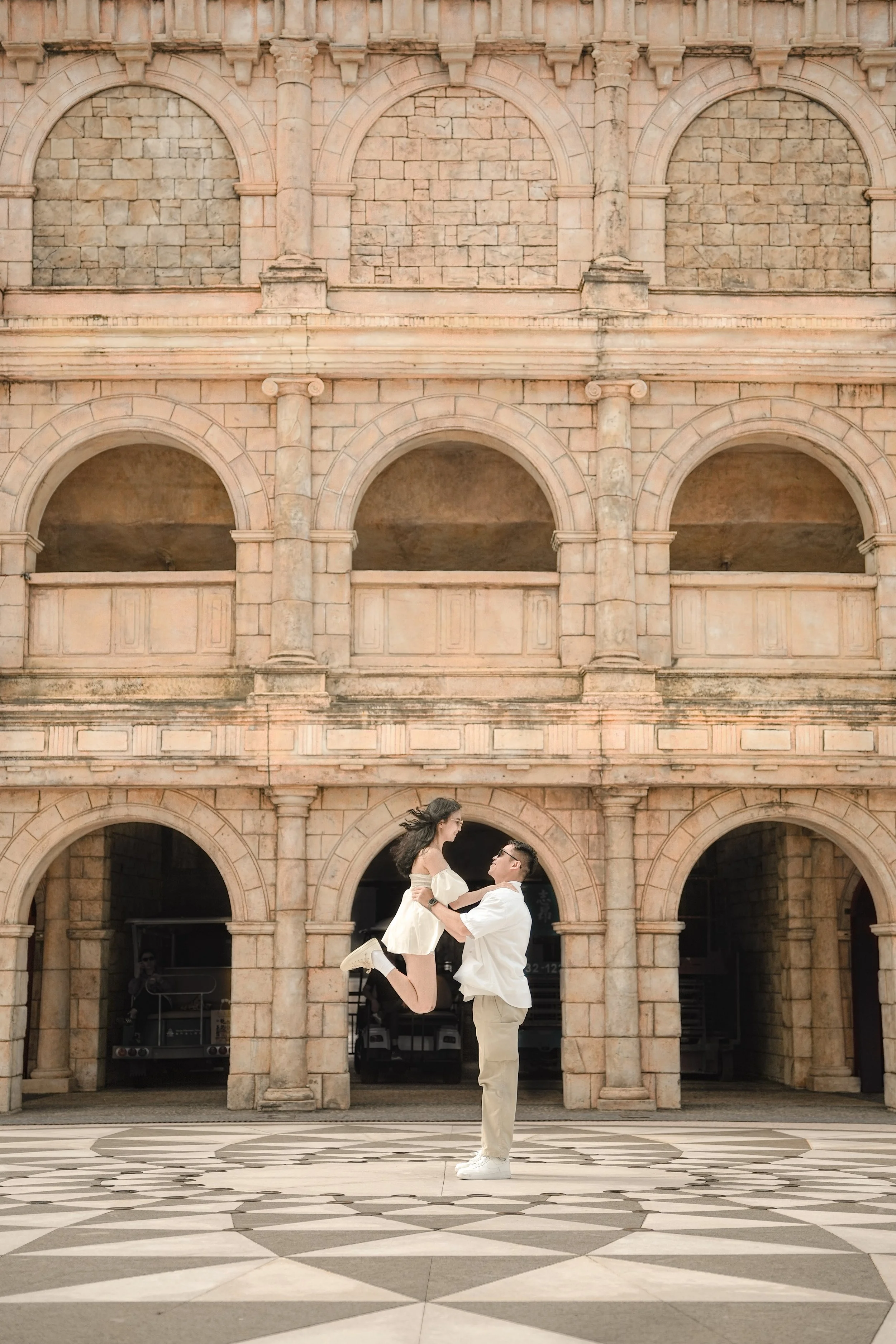 A man lifting a woman in the air in front of a historic stone building with arched windows.