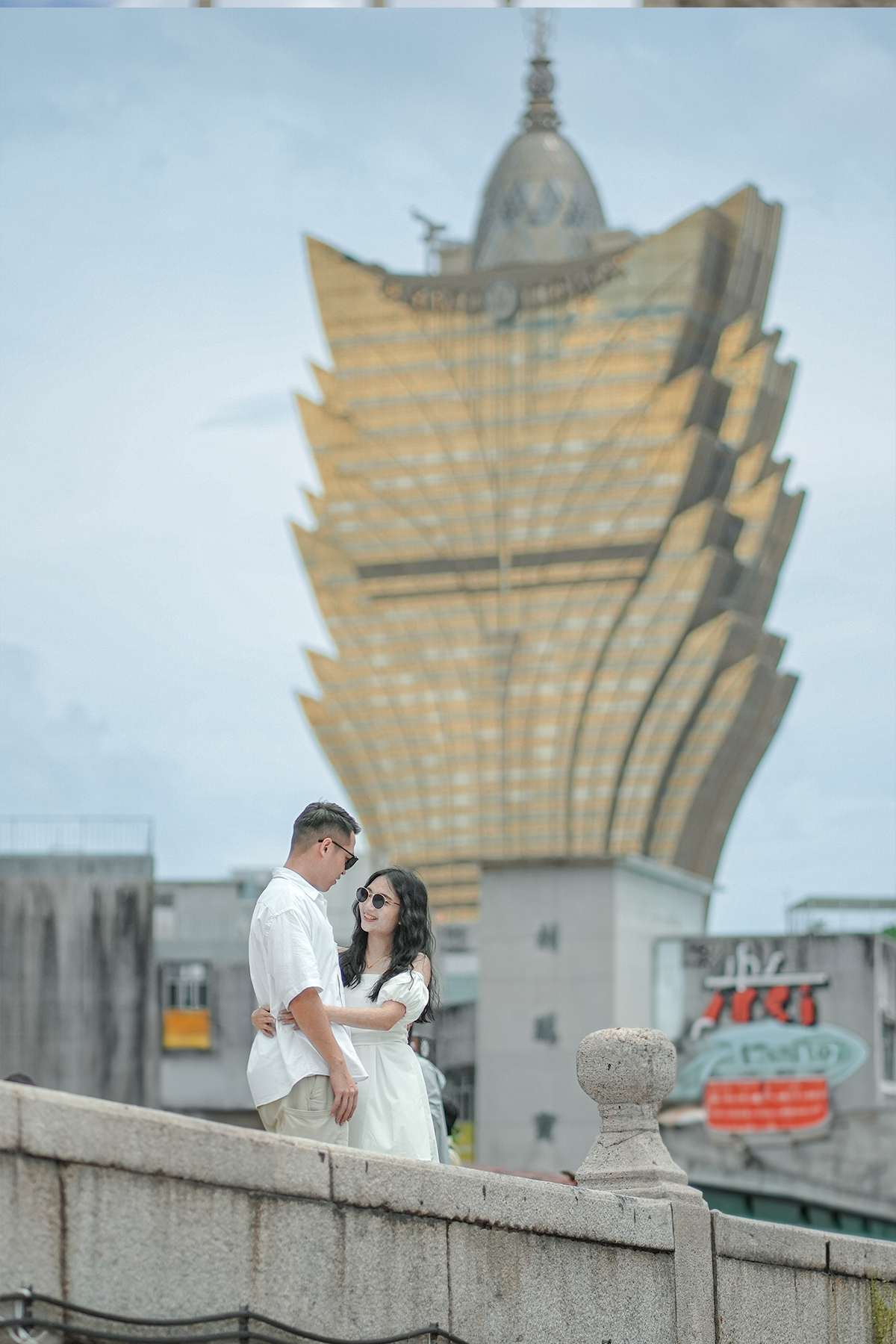 A young couple standing on a stone ledge, smiling at each other, with a modern, unique golden high-rise building in the background.