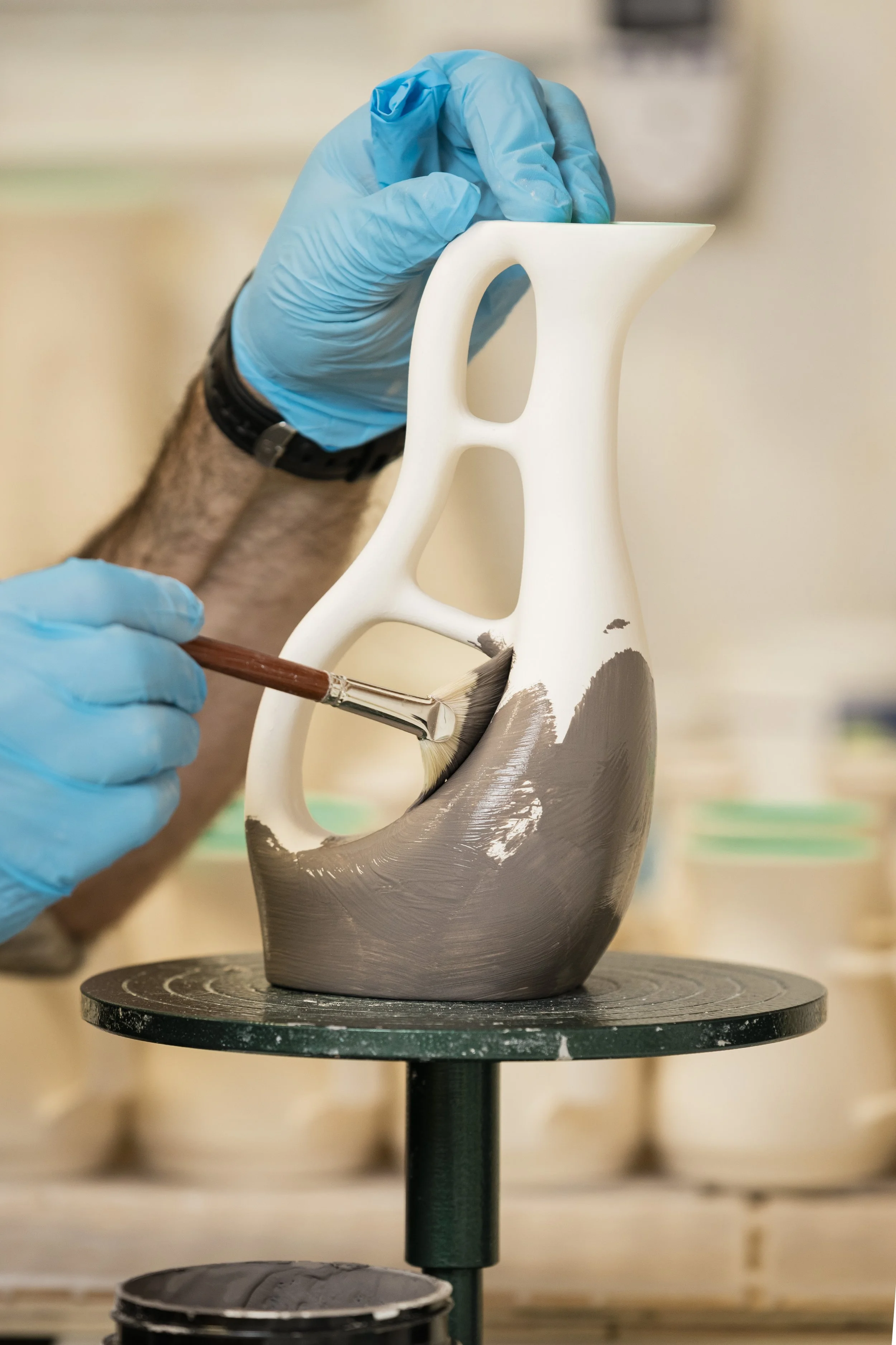 A person wearing blue gloves is painting a decorative ceramic vase with a brush in a workshop.