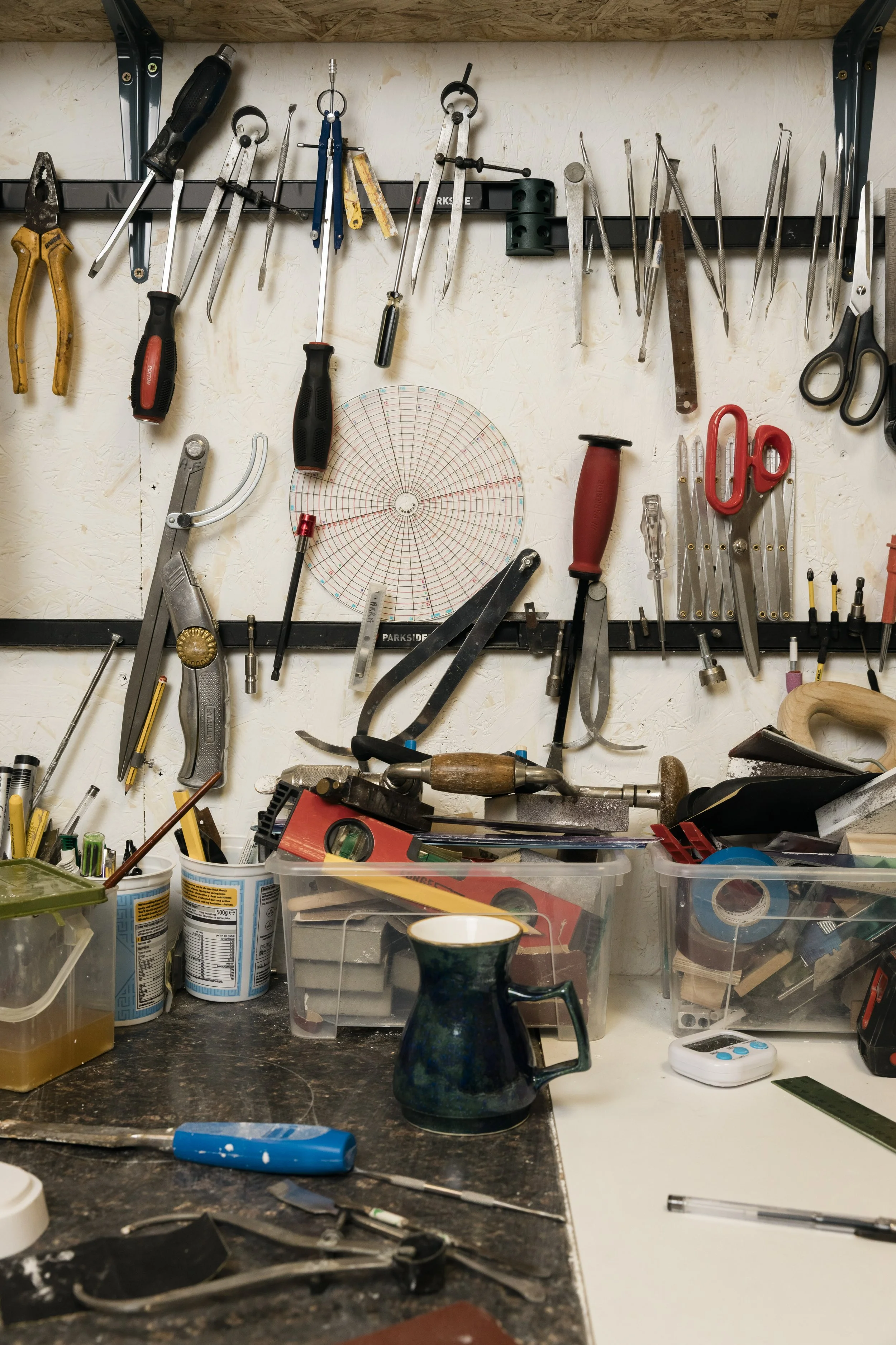 A ceramic studio with tools hanging on the wall, containers with supplies, a ceramic pitcher on the workbench, and various small tools and items scattered across the surface.