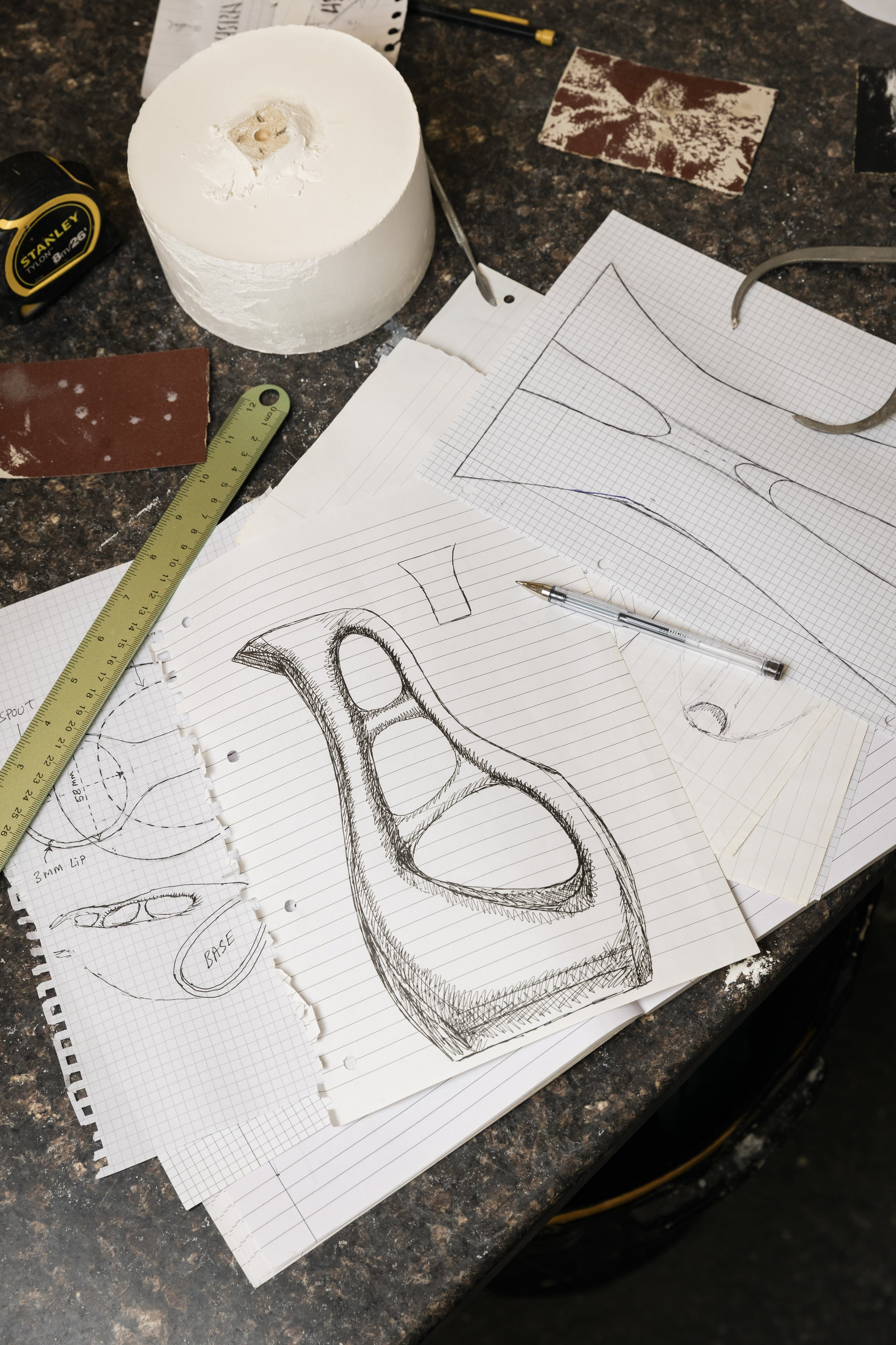 Sketches and plans of a ceramic design, drawing tools, ruler, and materials organized on a dark granite work surface.