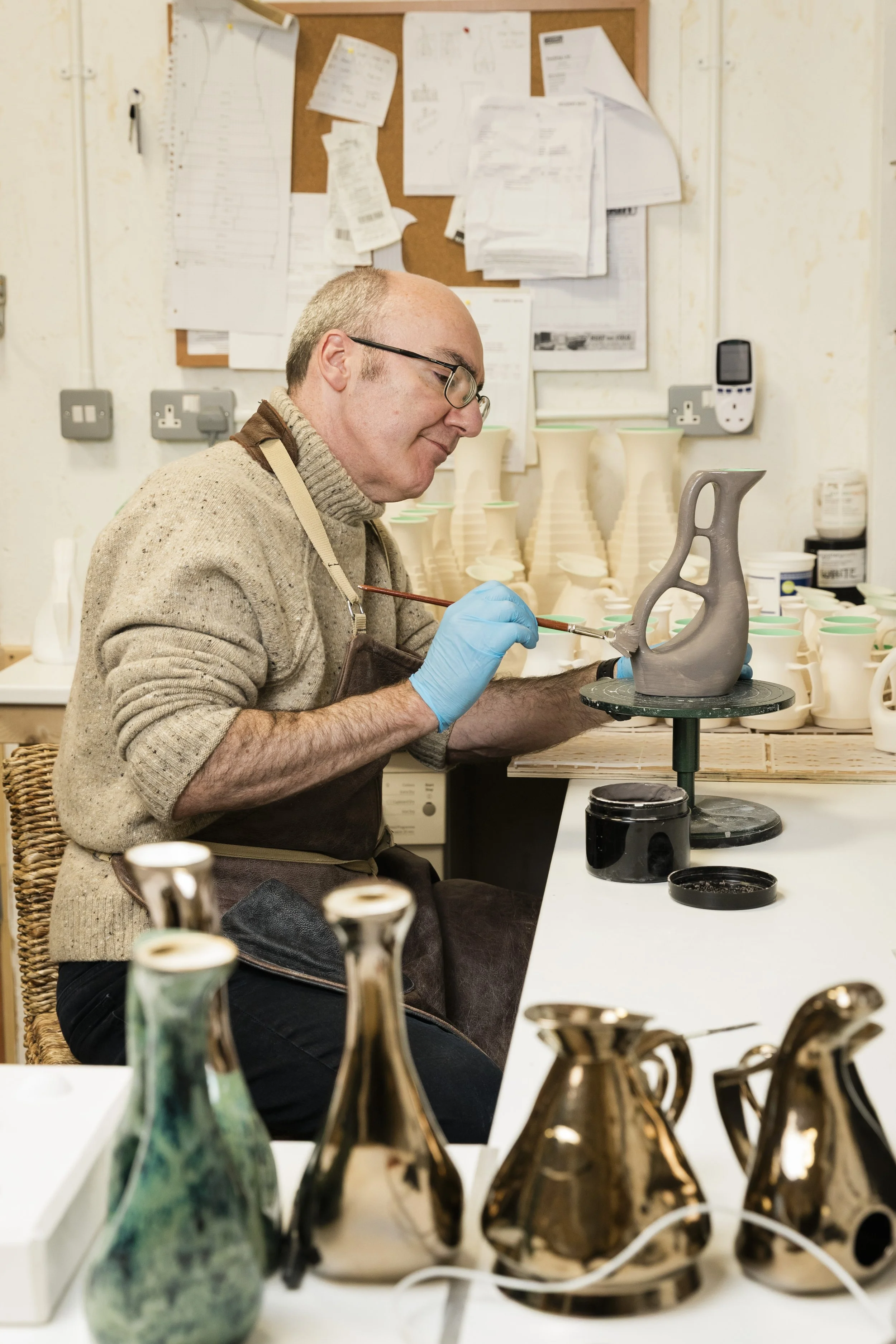 Scottish ceramicist, Arra Fletcher, painting a ceramic vase in his studio with various vases and ceramic items around.