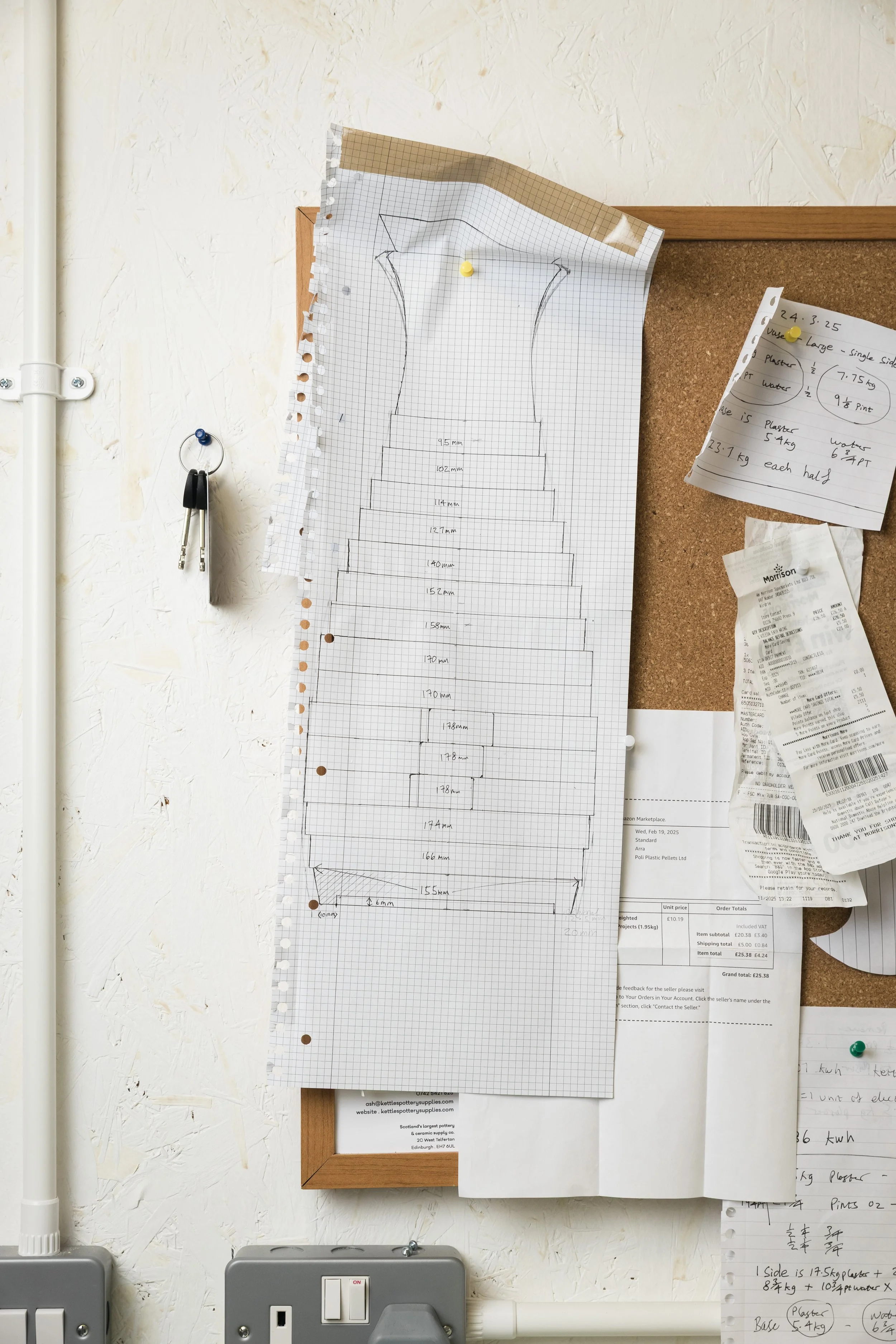 A large sheet of graph paper with a design and measurements pinned to a corkboard on a wall, with various papers and receipts nearby.