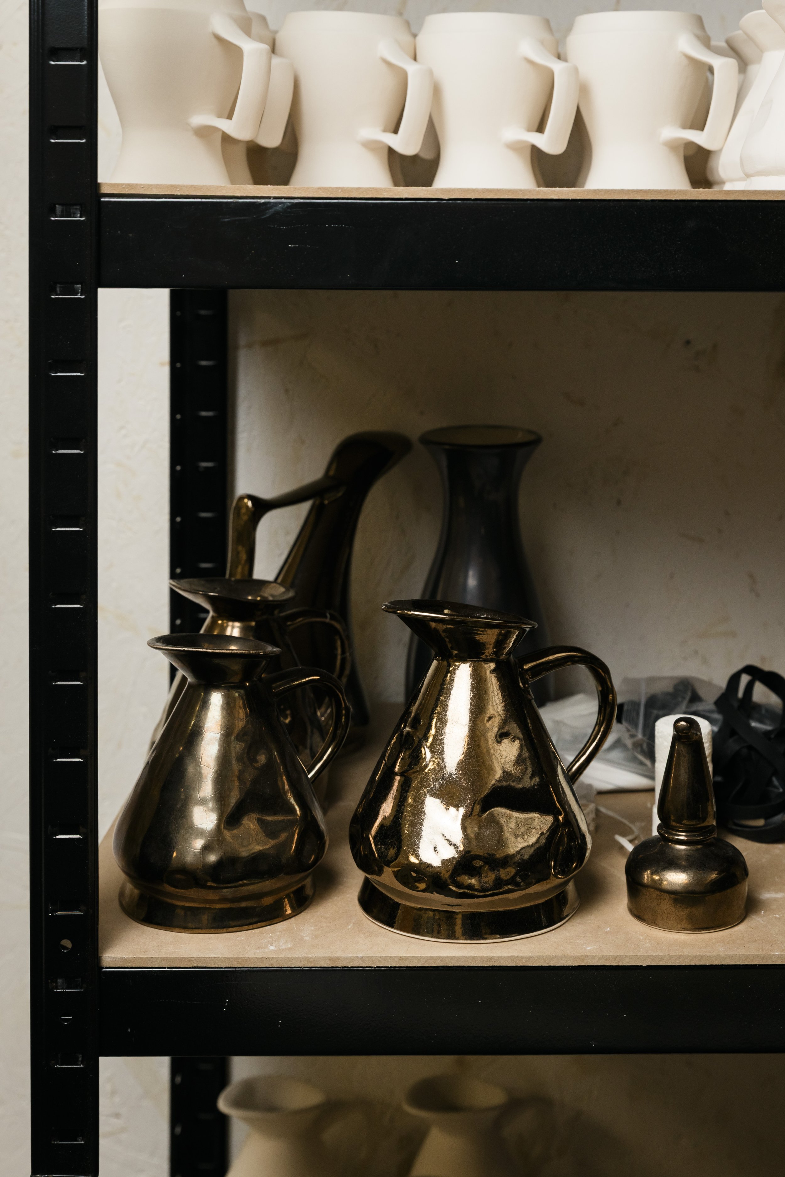 Shelf with shiny gold ceramic vases and pitchers, and white ceramic cups on the top shelf.