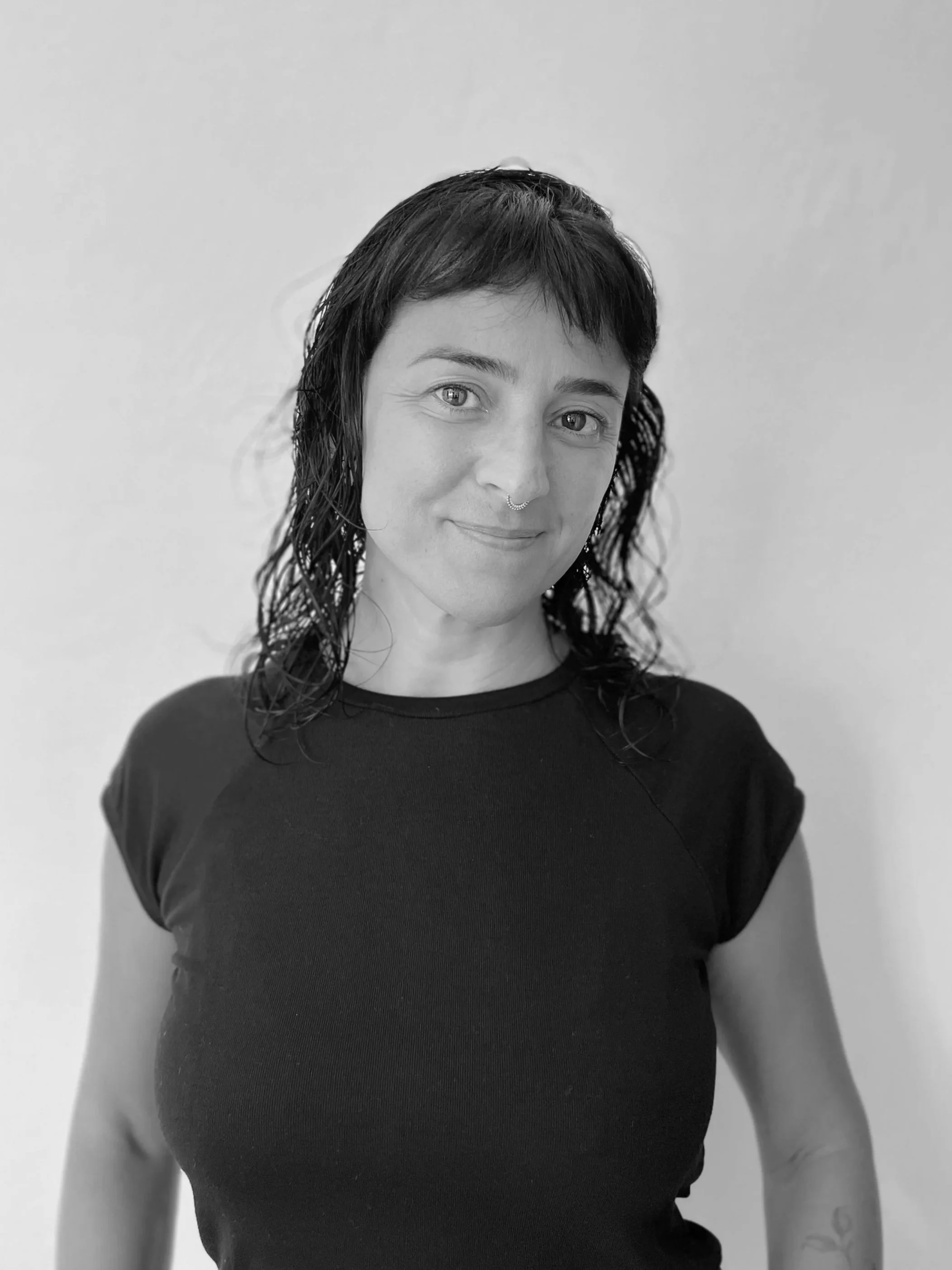 Retrato de una mujer con cabello rizado y oscuro, usando una camiseta negra, sonriendo suavemente hacia la cámara, en un fondo blanco.