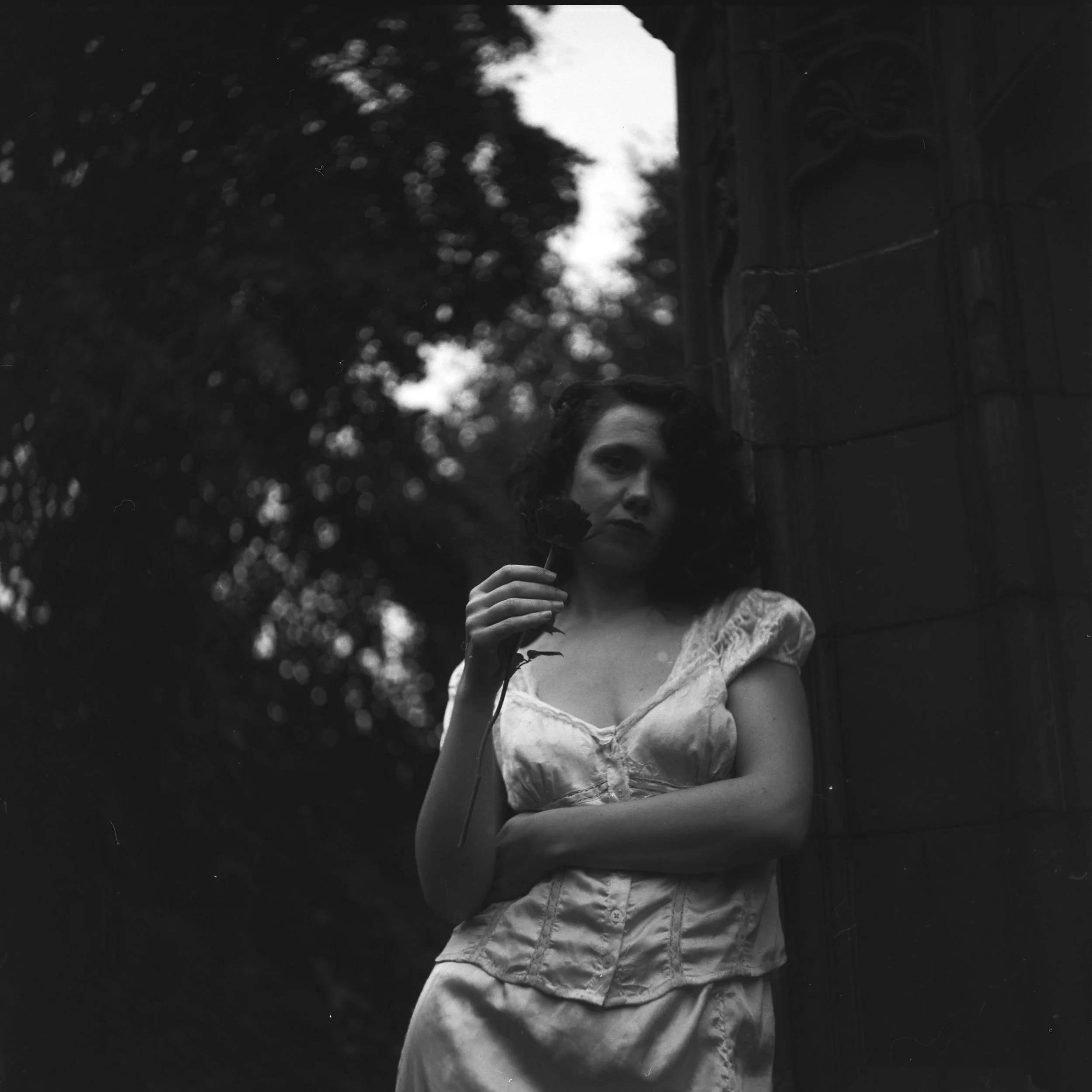 Black and white photograph of a woman with dark hair holding a small branch with leaves, standing near an ornate stone structure outdoors with blurred trees in the background.