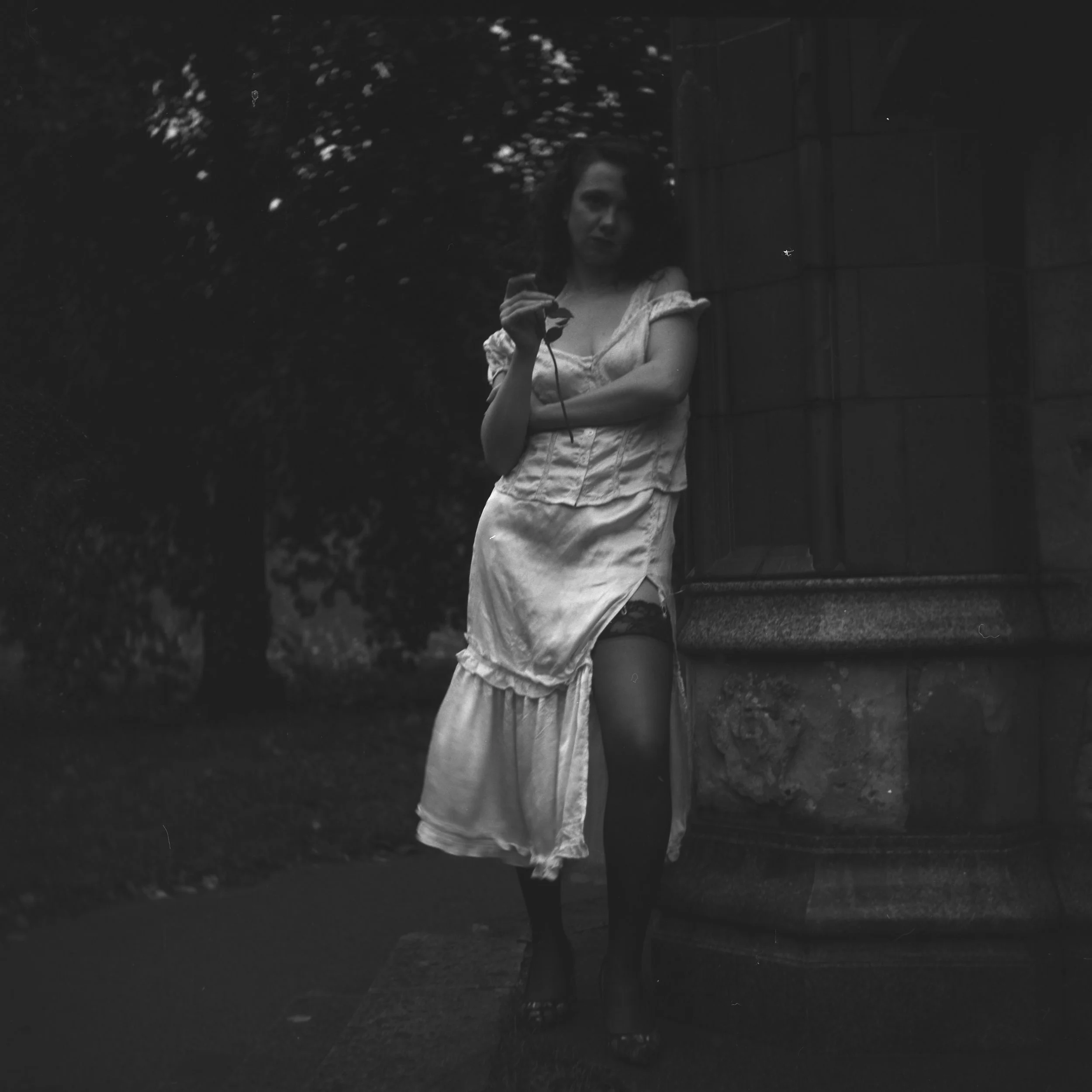 A woman in a vintage white dress standing outside near a wall and some bushes at night, holding a pair of sunglasses.