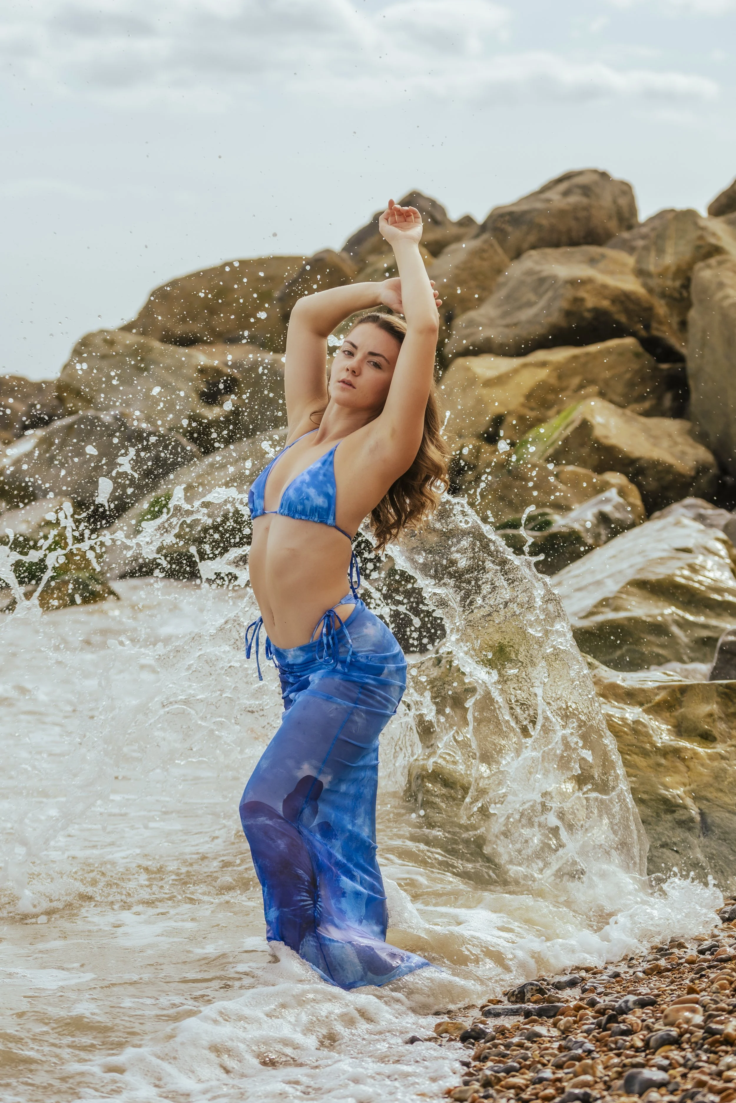 Woman in a blue bikini and sheer blue pants posing in shallow ocean water near rocks, with waves splashing around.