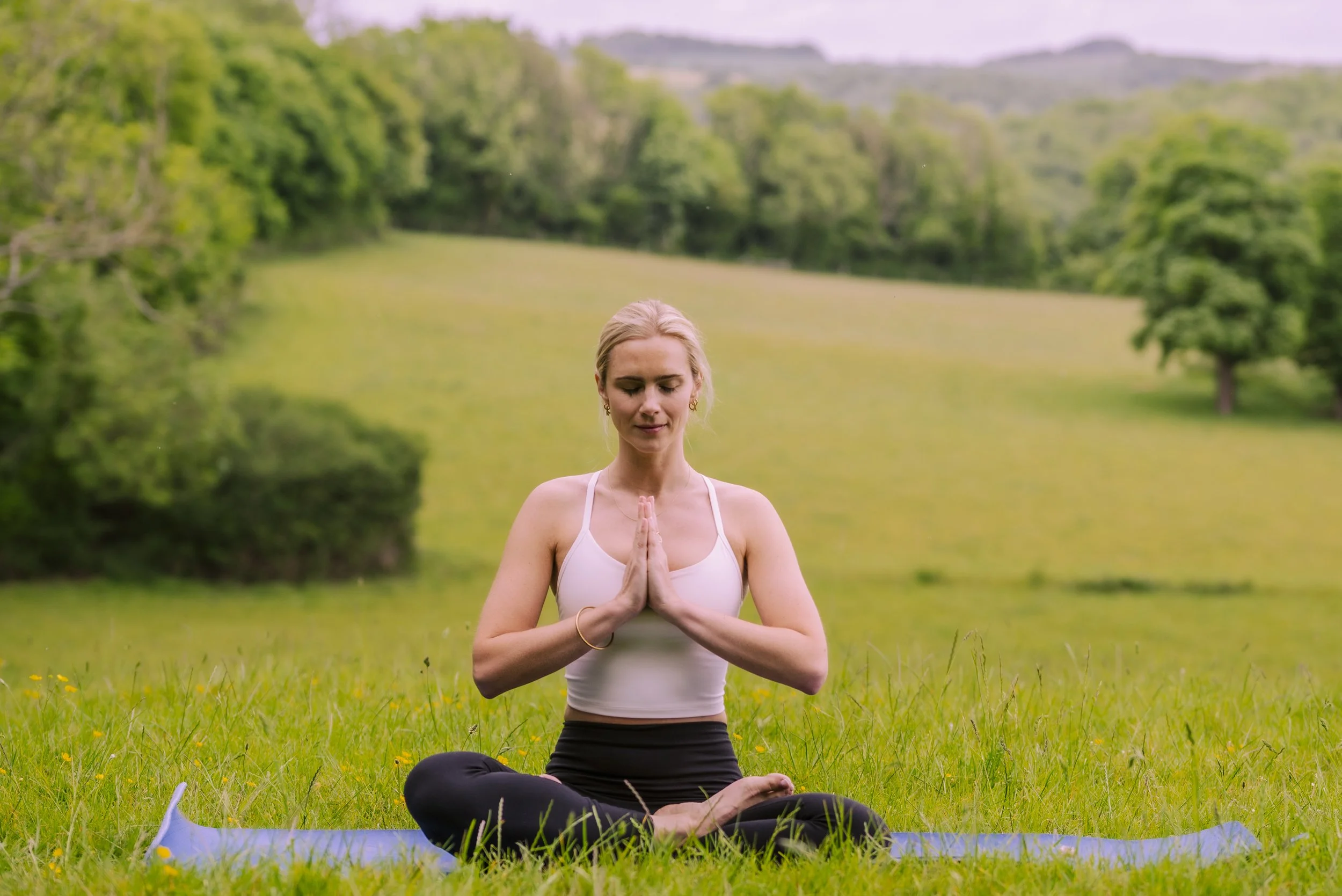 A woman practicing yoga outdoors in a grassy field, sitting in a cross-legged position with hands in prayer pose, eyes closed, surrounded by trees and hills in the background.