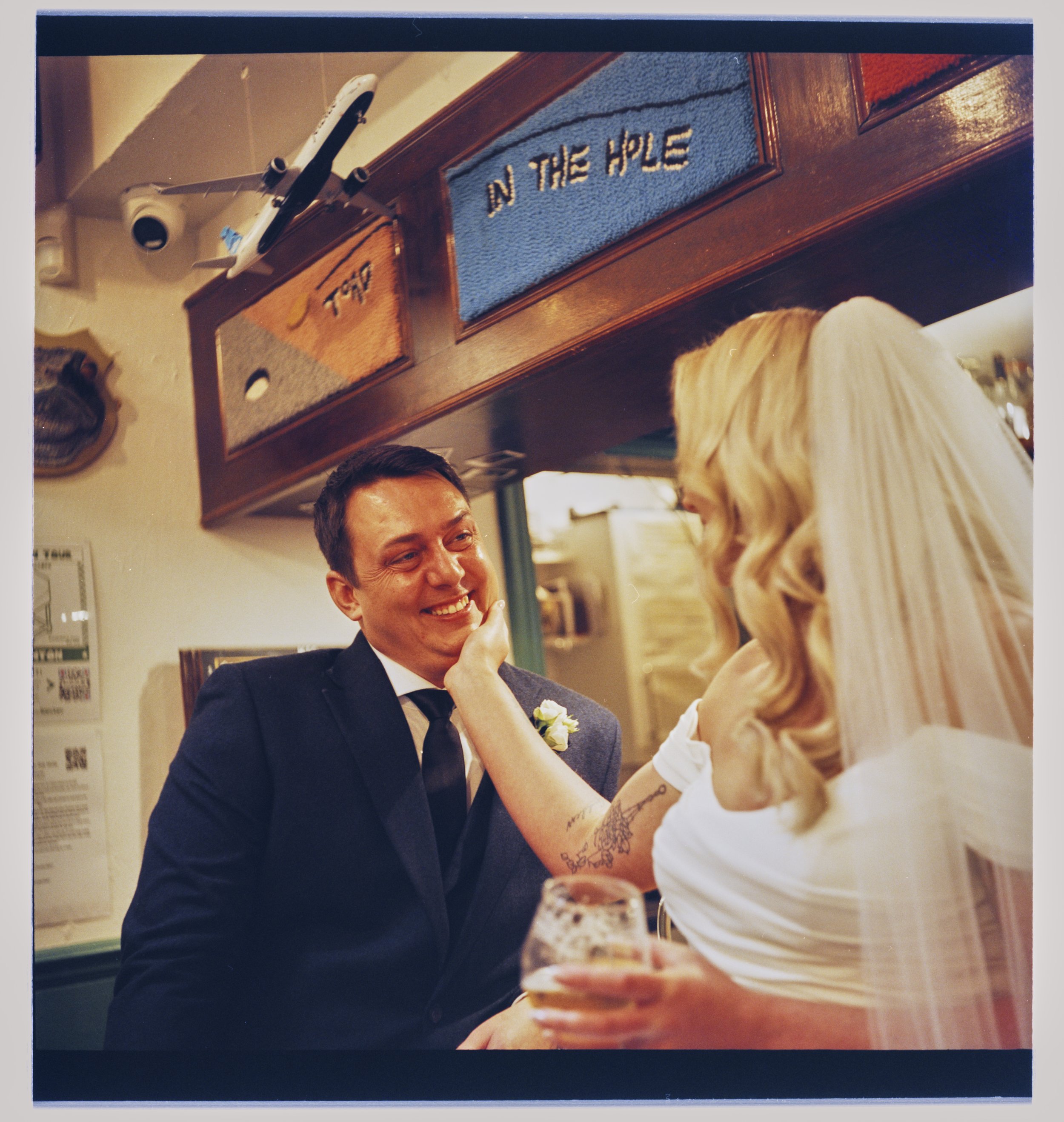 A man and woman are sitting at a table in a restaurant, smiling and talking. The man is wearing a dark suit and tie, and the woman has blonde hair and is dressed in white. The woman has a tattoo on her arm, and she is holding a drink. There are two r