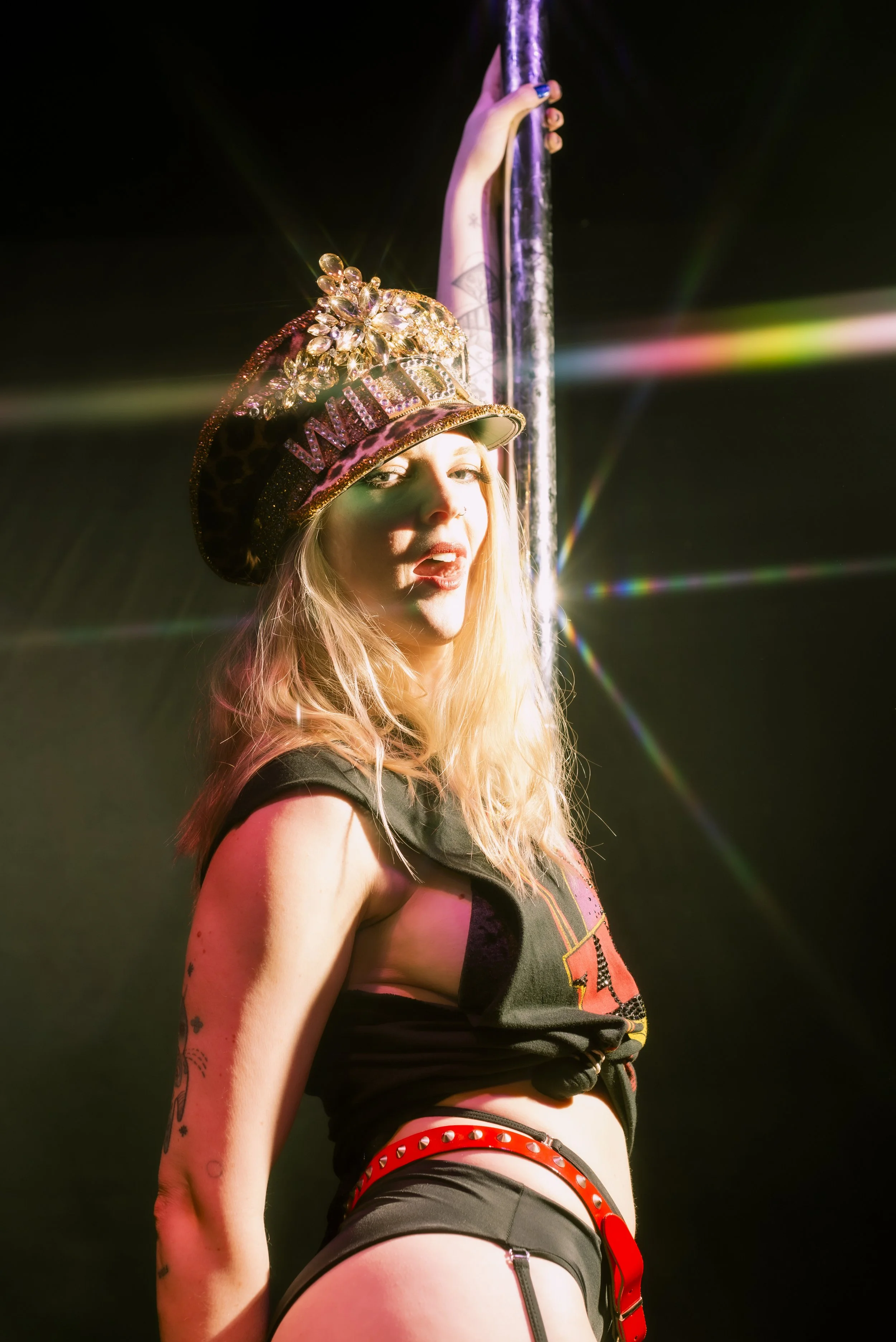 A woman with long blonde hair wearing a black crop top, black shorts with red studded belts, a leopard print cap decorated with jewelry, and holding onto a pole with her right hand. She appears to be performing a pole dance.