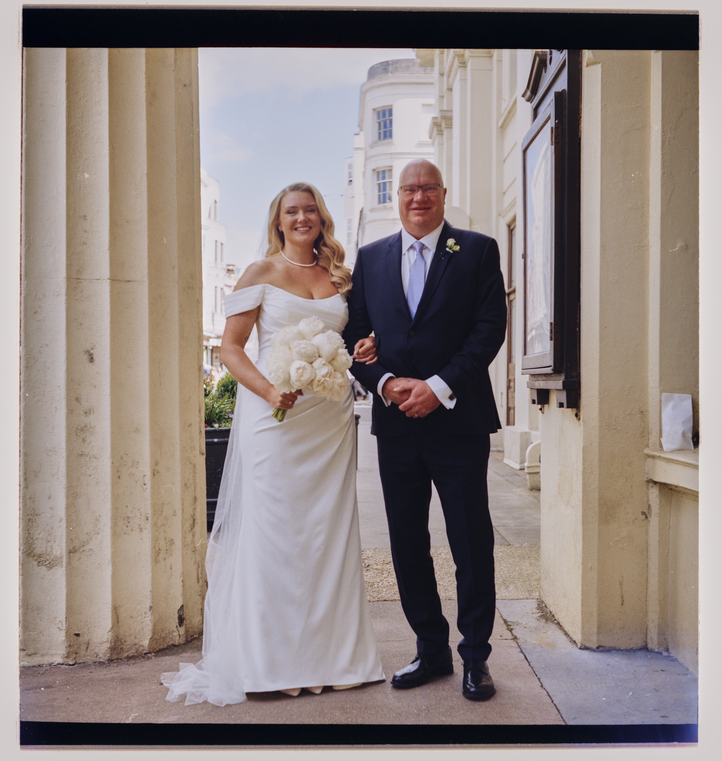 A bride in a white off-shoulder wedding gown holding a bouquet of white flowers, standing next to a smiling groom in a dark suit, outside a building with classical architecture.