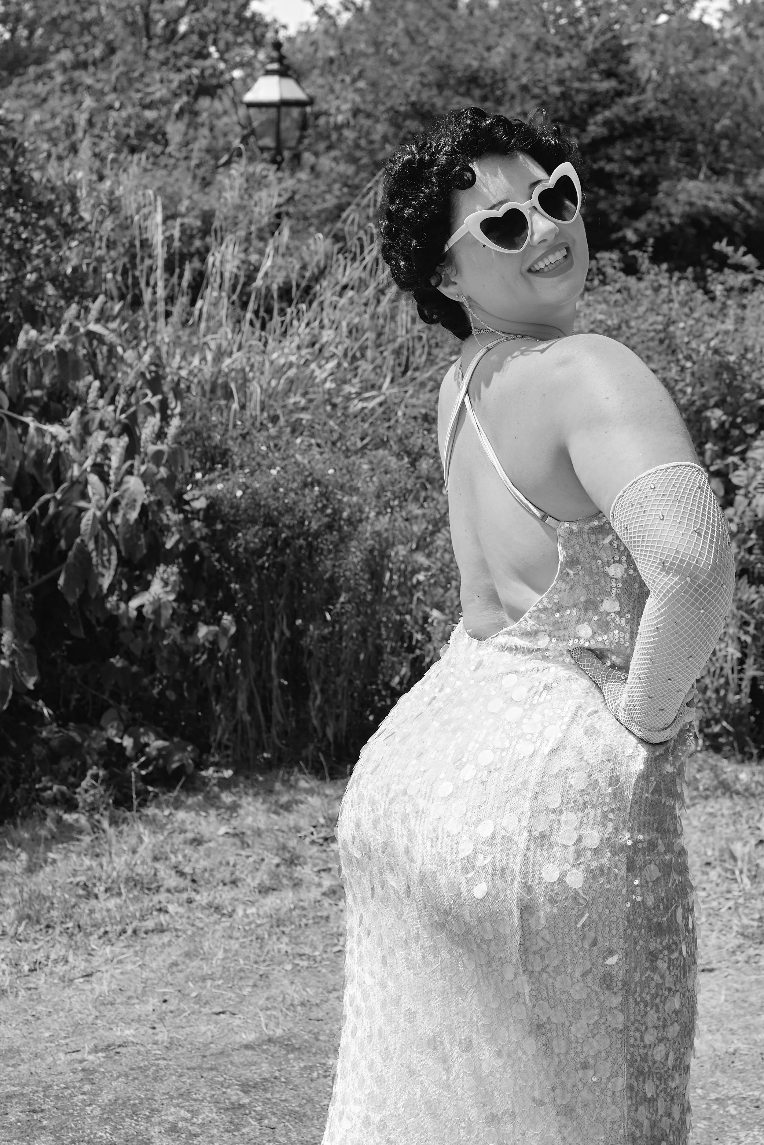 A woman in a vintage style dress with sequins, wearing heart-shaped sunglasses, standing outdoors with a natural background, smiling and looking over her shoulder.
