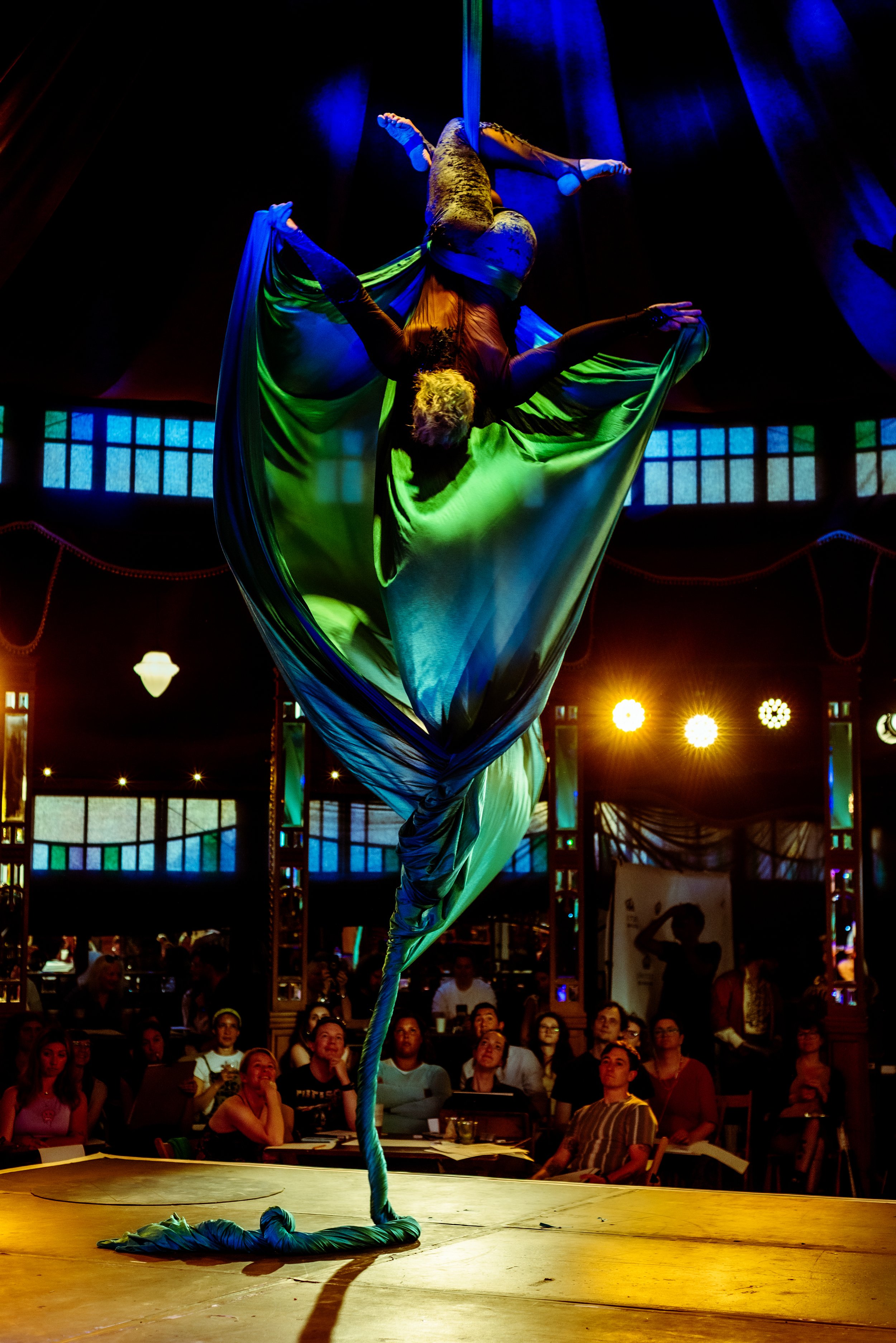 Aerial silk performer hanging upside down on green silk fabric with an audience watching in a dimly lit indoor venue with colorful lighting.