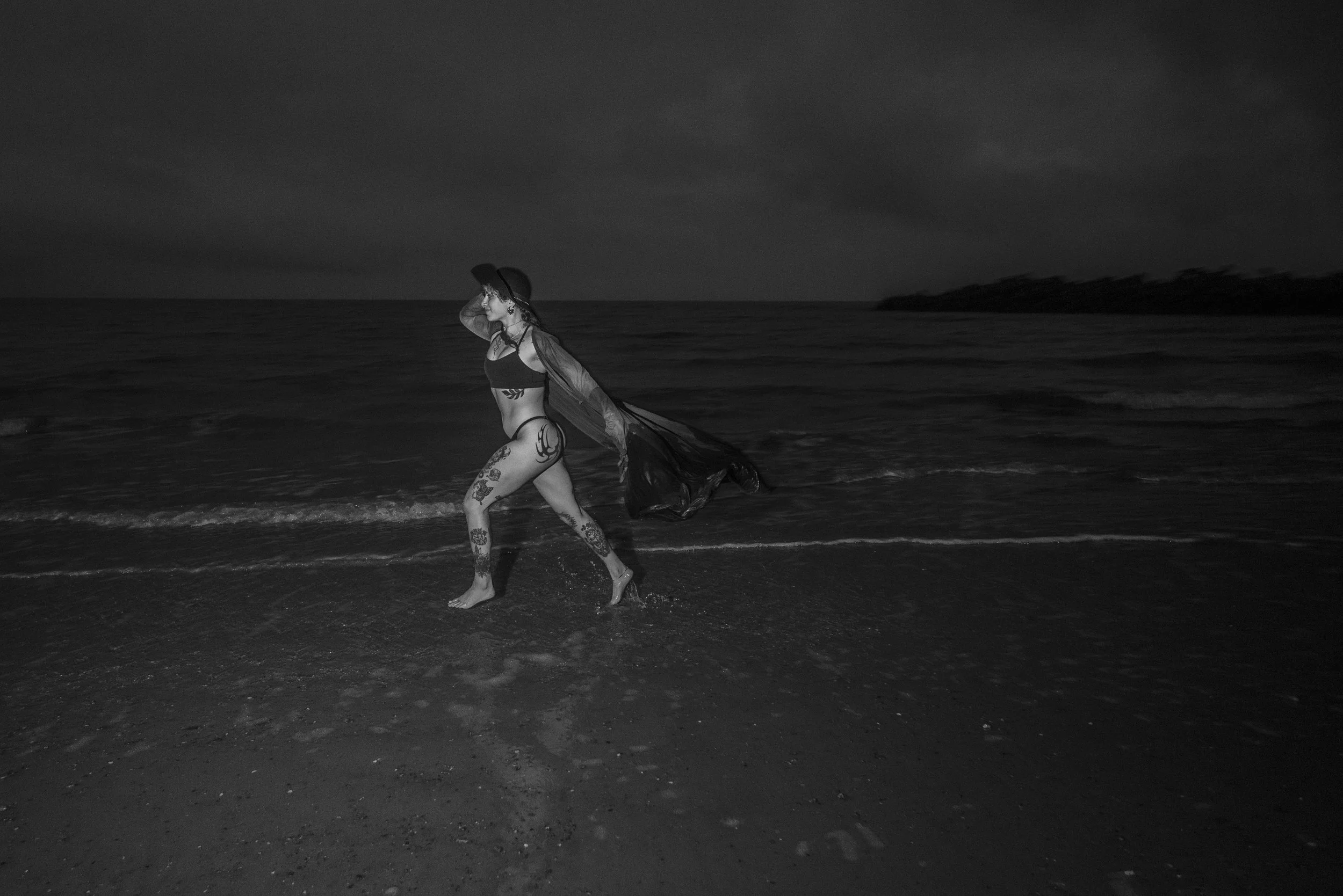 A black-and-white photo of a woman with tattoos walking by the ocean at dusk or night. She is wearing a hat, a crop top, and a sheer cover-up, with water around her feet.